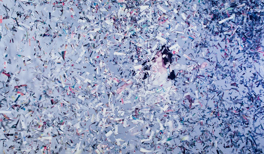 Performer suspended amid swirling shredded newspaper in a staged scene evoking a human caught in extreme weather.