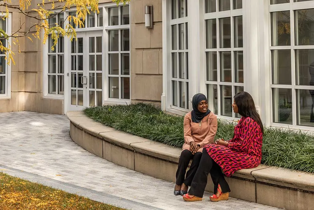 Two women of color sitting on a curved stone bench outside of Shad hall, engaged in a warm conversation