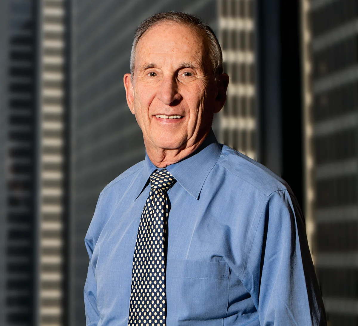 Person wearing a blue dress shirt and patterned tie, standing in front of tall office buildings in sunlight.