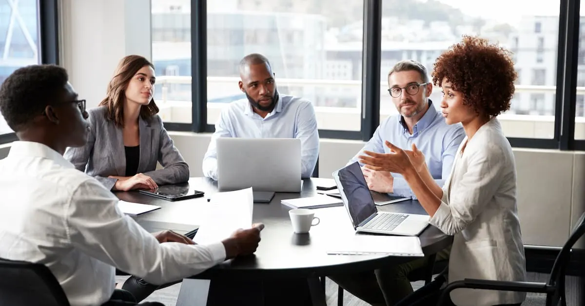 A group of five co-workers sitting around a table in a bright office, discussing work with laptops and documents.