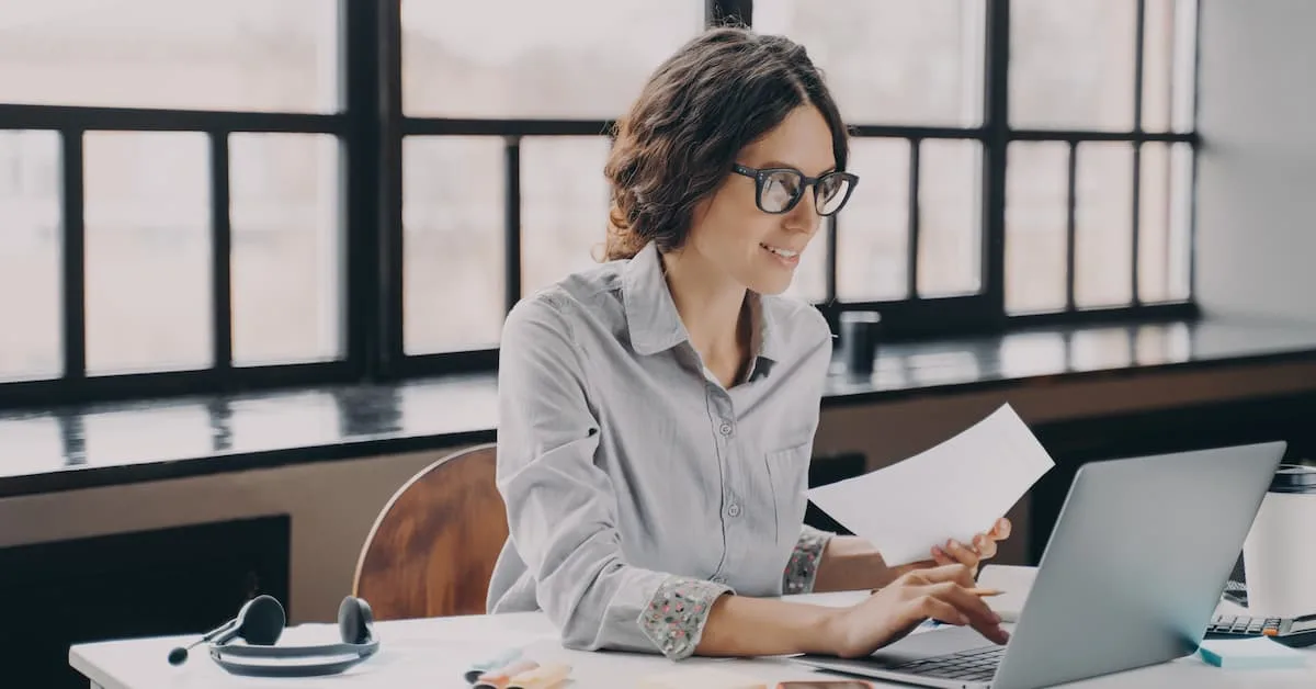 Woman reviewing documents while working on a laptop at a desk.