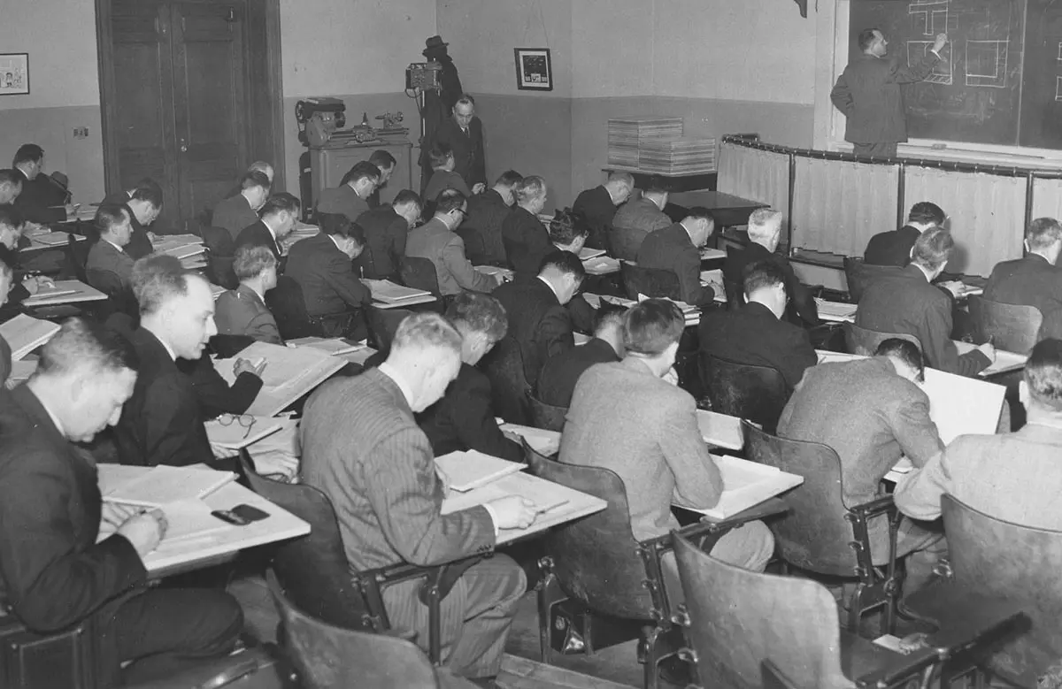 Black and white photograph of a classroom of men taking notes while a male professor writes on a chalkboard.