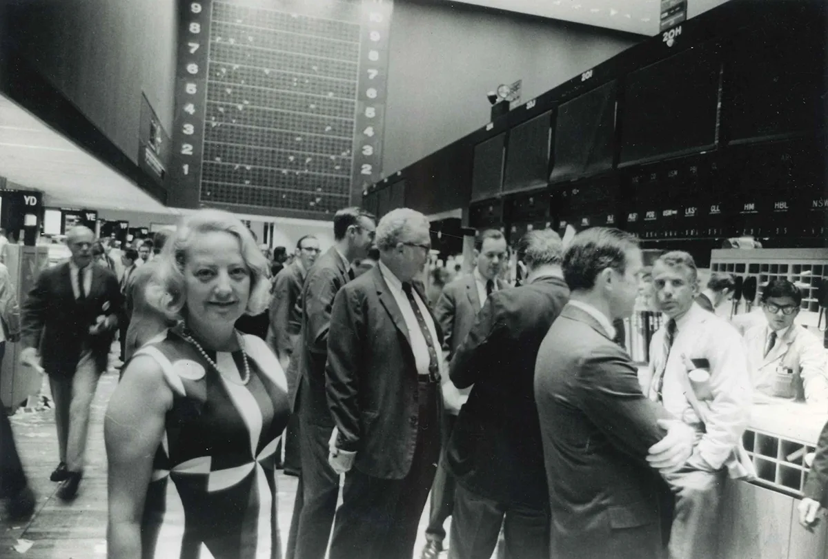 Woman in print dress standing on Stock Exchange floor surrounded by men