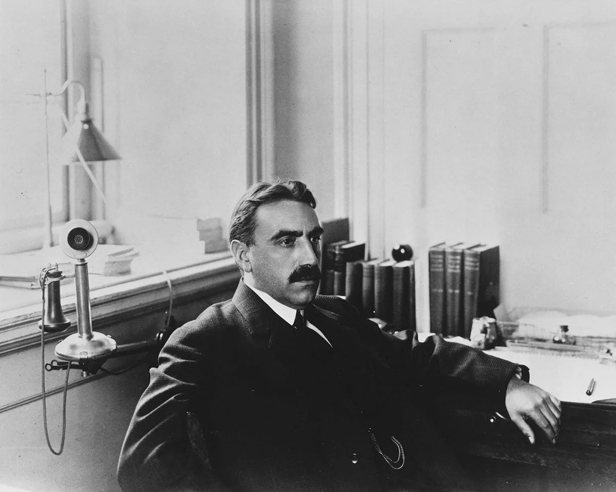 Dean Edwin F. Gay sits in an office, gazing to the right. Behind him sits an old telephone. Across his decks are upright books and scattered papers.