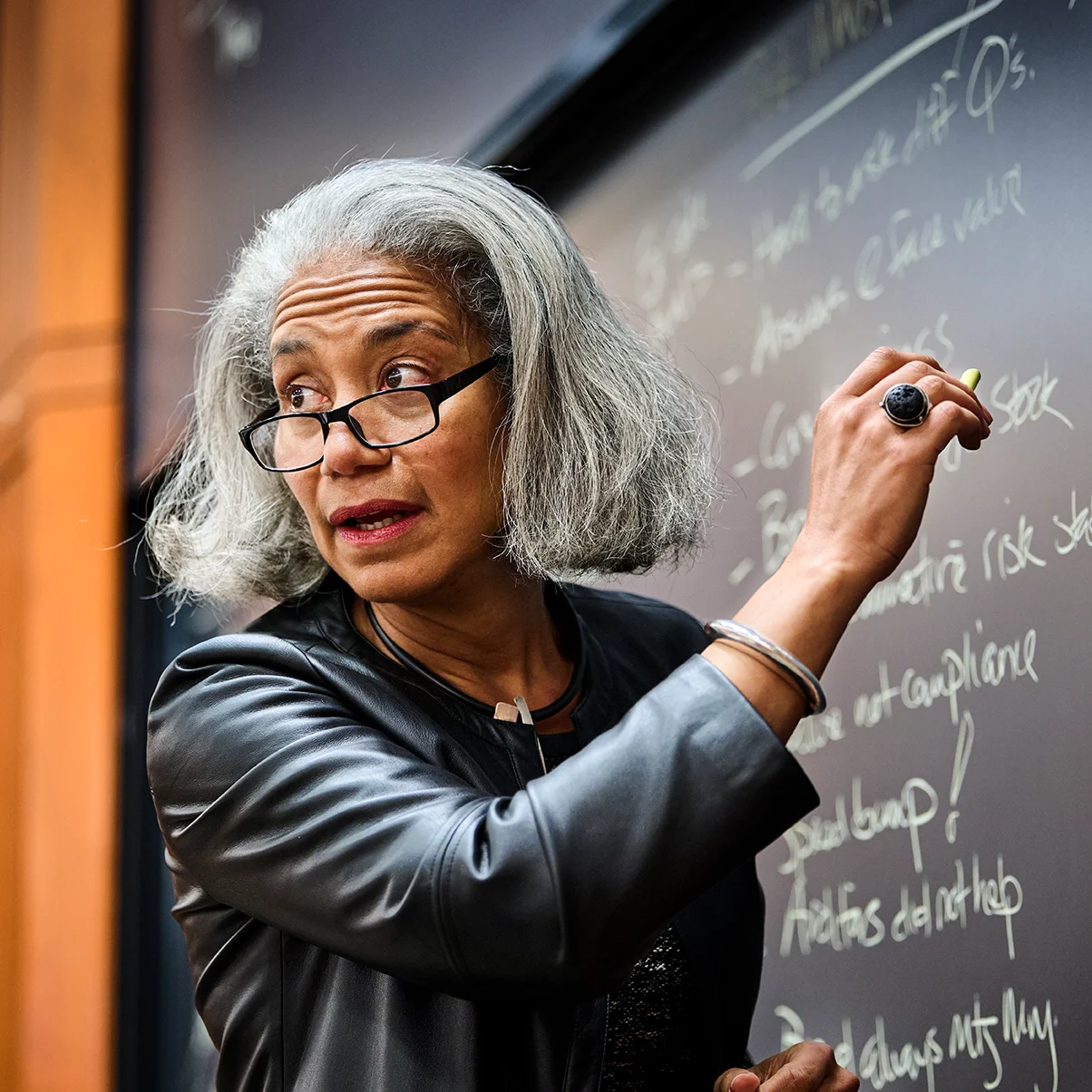 A confident professor with silver hair and glasses writes on a chalkboard, engaged in an animated lecture in a classroom setting