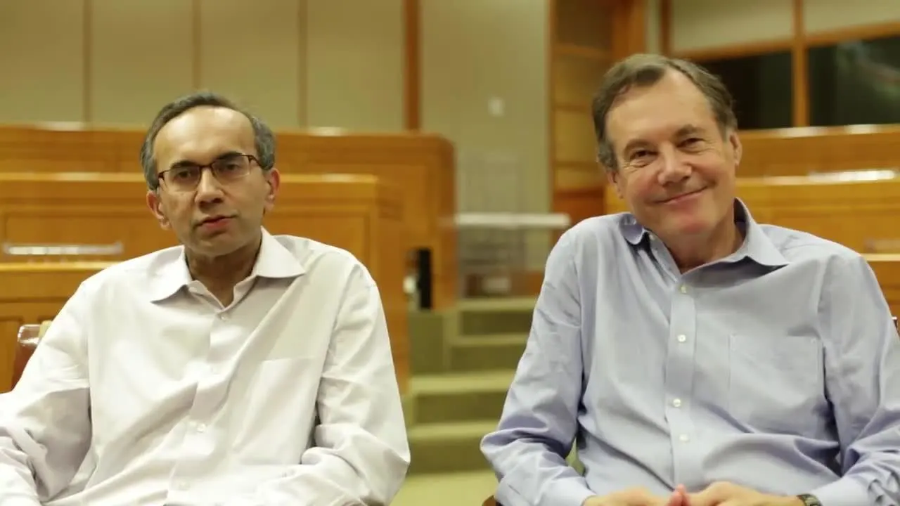 Two people seated in a lecture hall or auditorium, facing the camera, wearing light-colored button-down shirts.