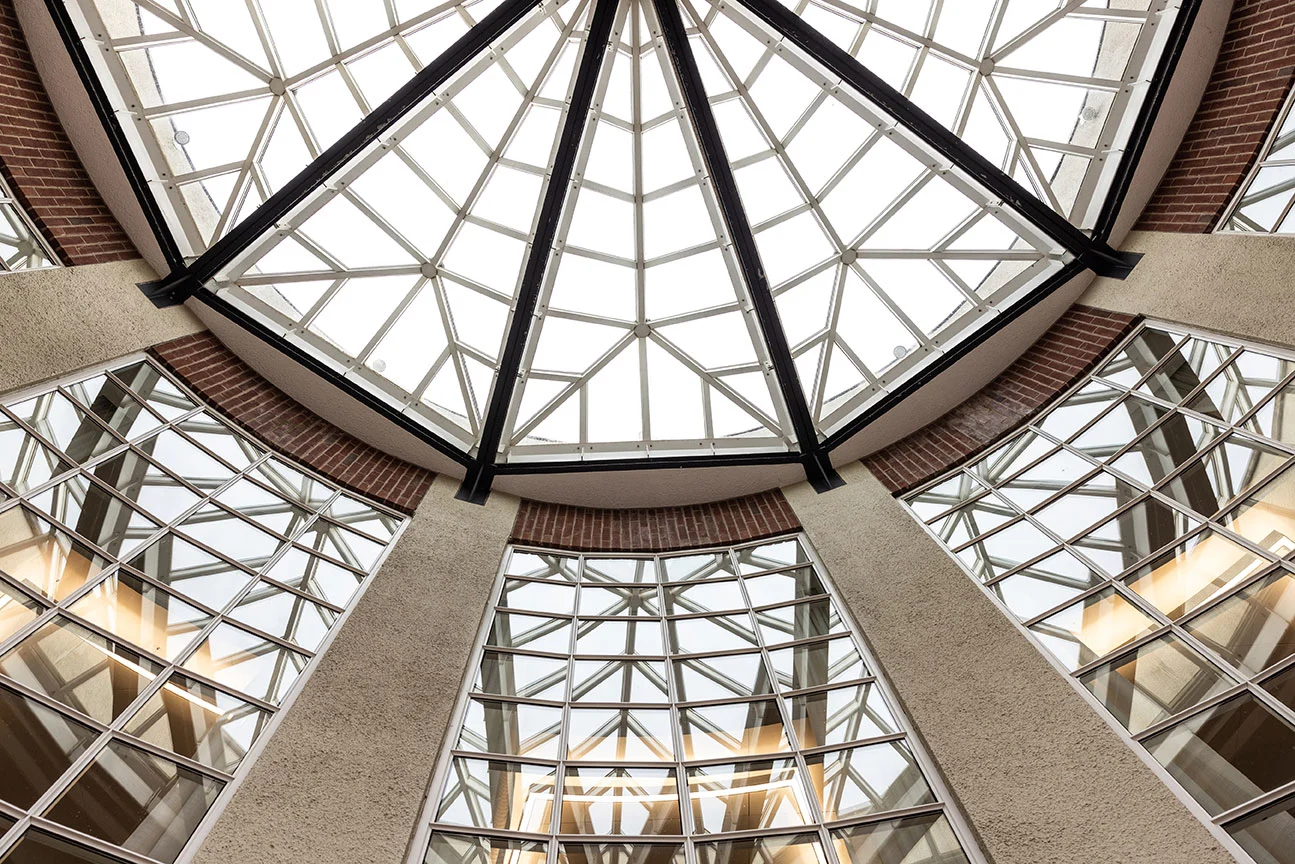 Upward view inside a modern atrium featuring a large geometric glass skylight with black steel beams, surrounded by tall reflective windows, brick accents, and textured concrete walls.