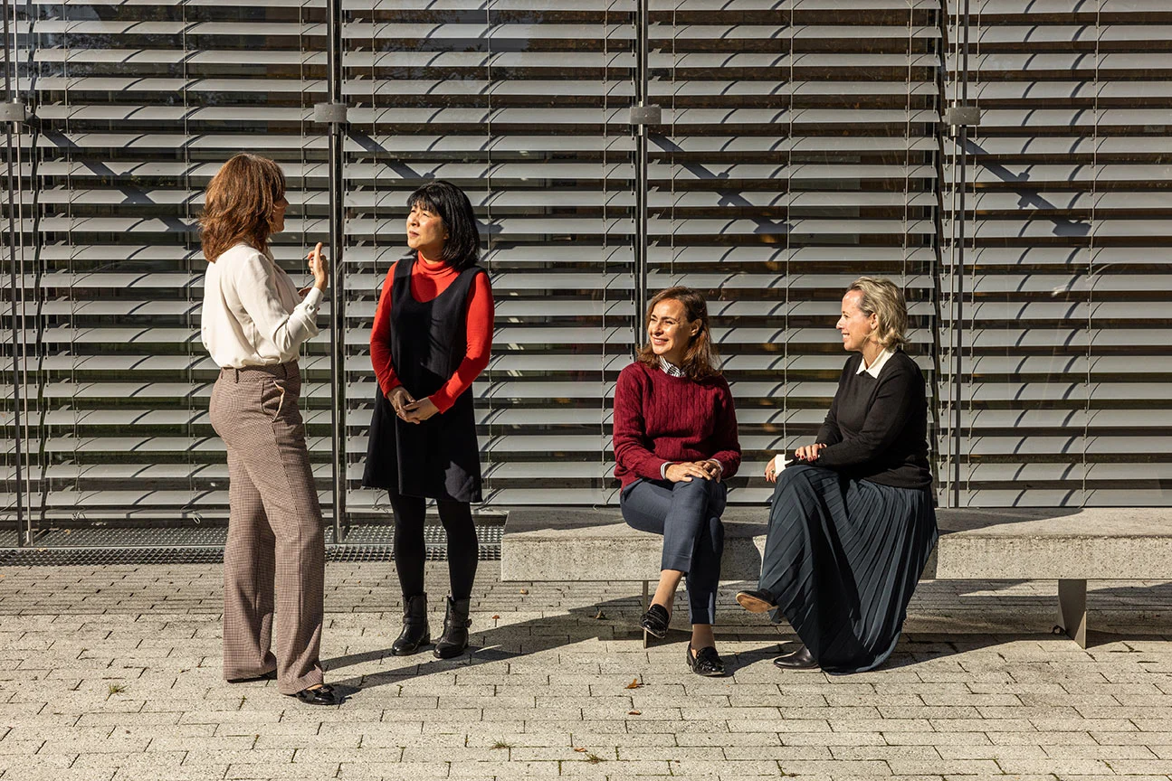 A group of four professionally dressed women engage in conversation outside a modern building with horizontal metal blinds in the background