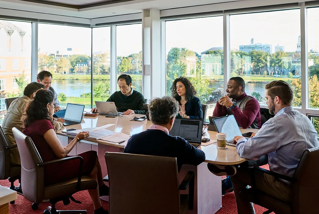 A group of executives sit at a round table and talk in a modern HBS office with large windows and Charles river view