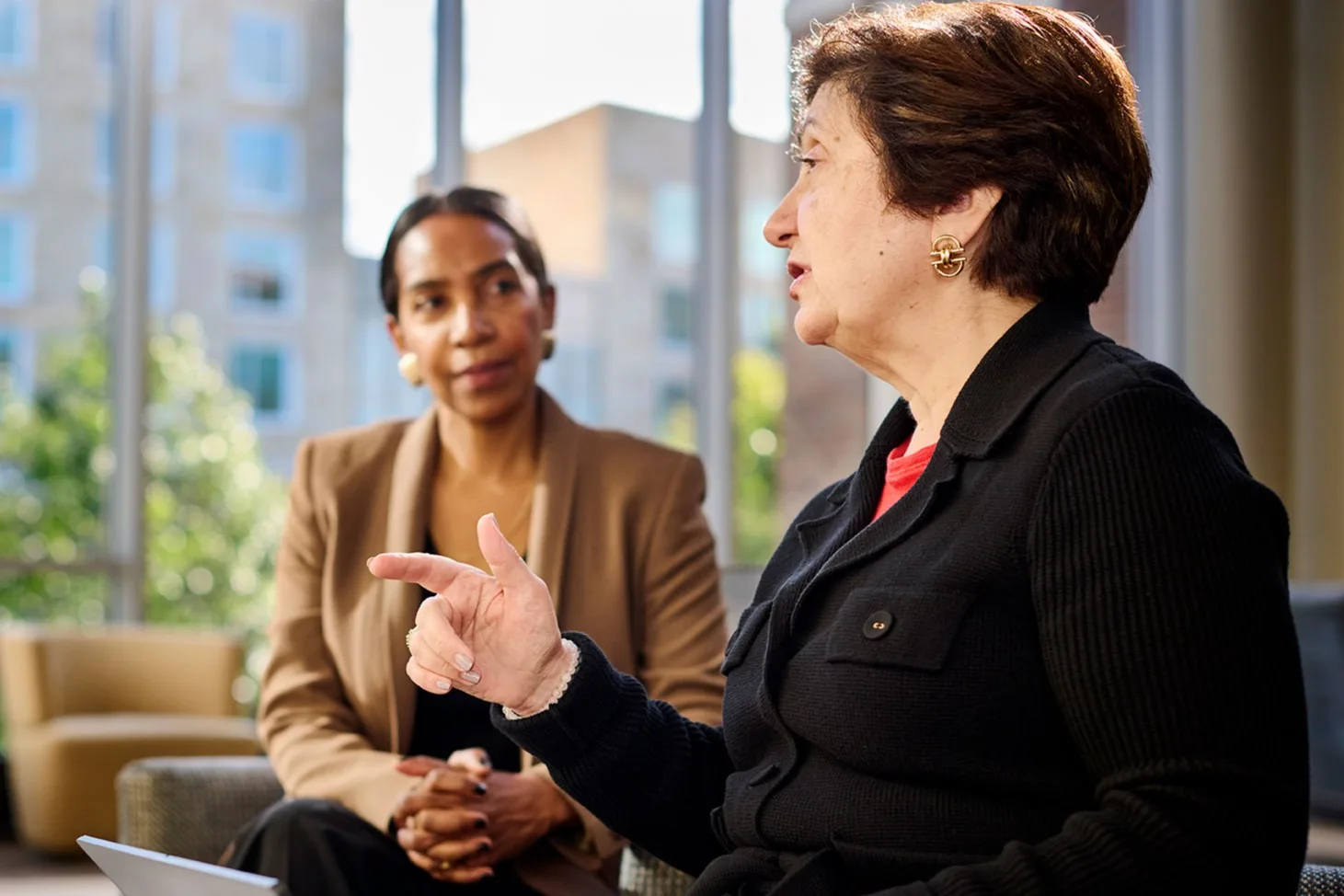Two female executives sit together, one with a laptop on her lap talking and the other listening intently
