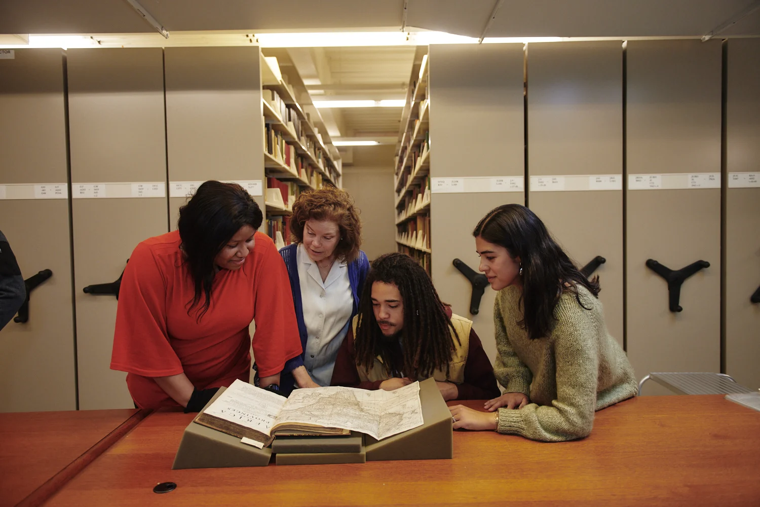 Four individuals gathered around a volume with books stacks in the background
