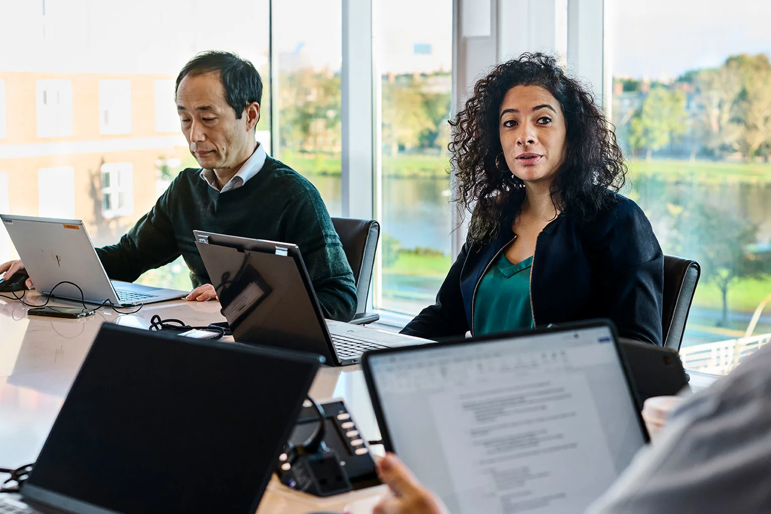 Participants sit and talk with their laptops open to review data in a conference room, overlooking HBS campus through large windows