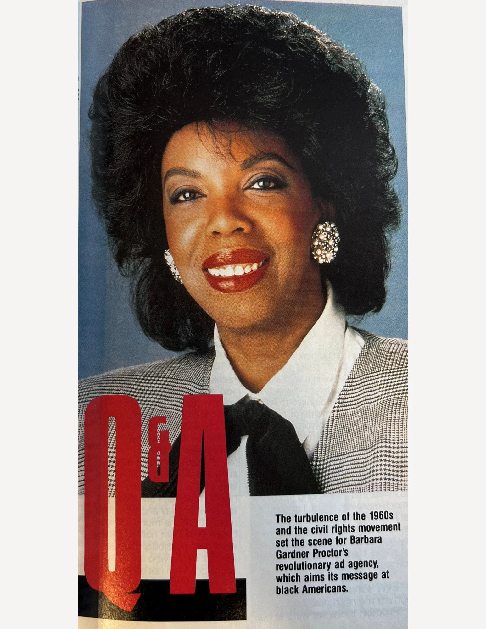 The face of a woman with large black hair, red lipstick, dark eye makeup, and big pearl-cluster earrings. She is smiling and wearing a black and white top with a black ascot.