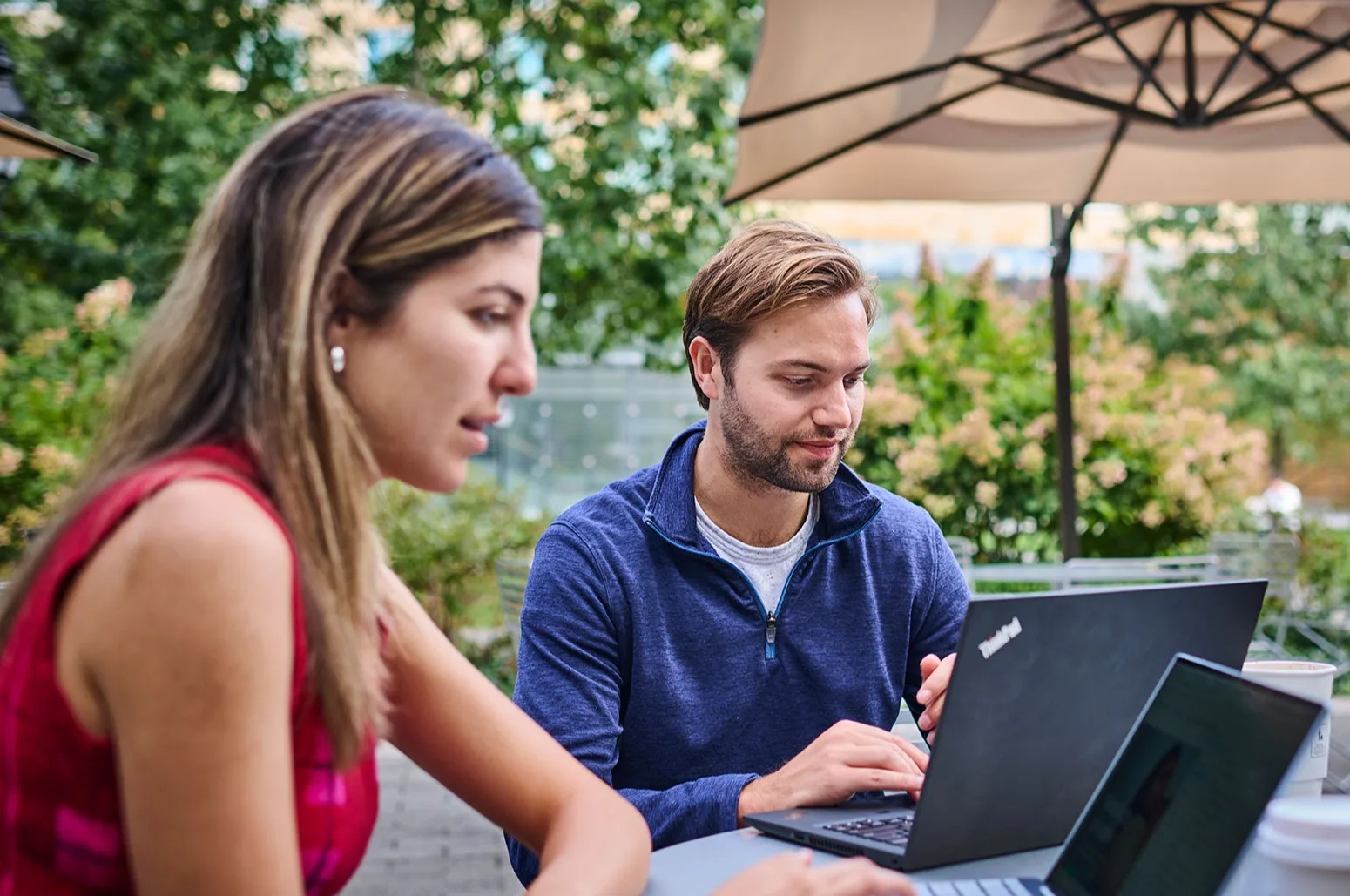 A female and a male participant sit outside at a table together while separately working on their laptops