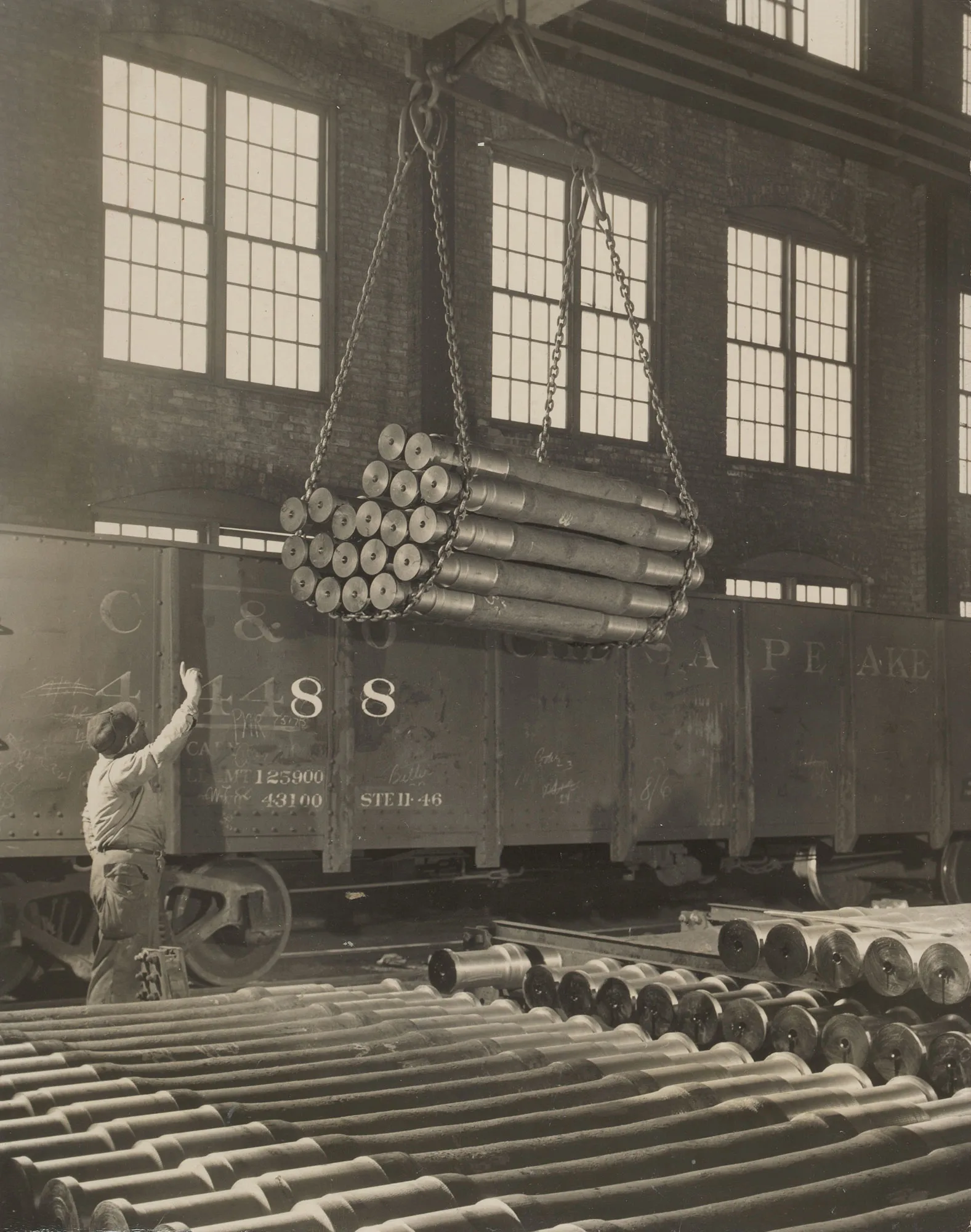 Worker signals crane as bundle of finished steel axles is loaded onto railcar for shipment at Gary Steel Works.