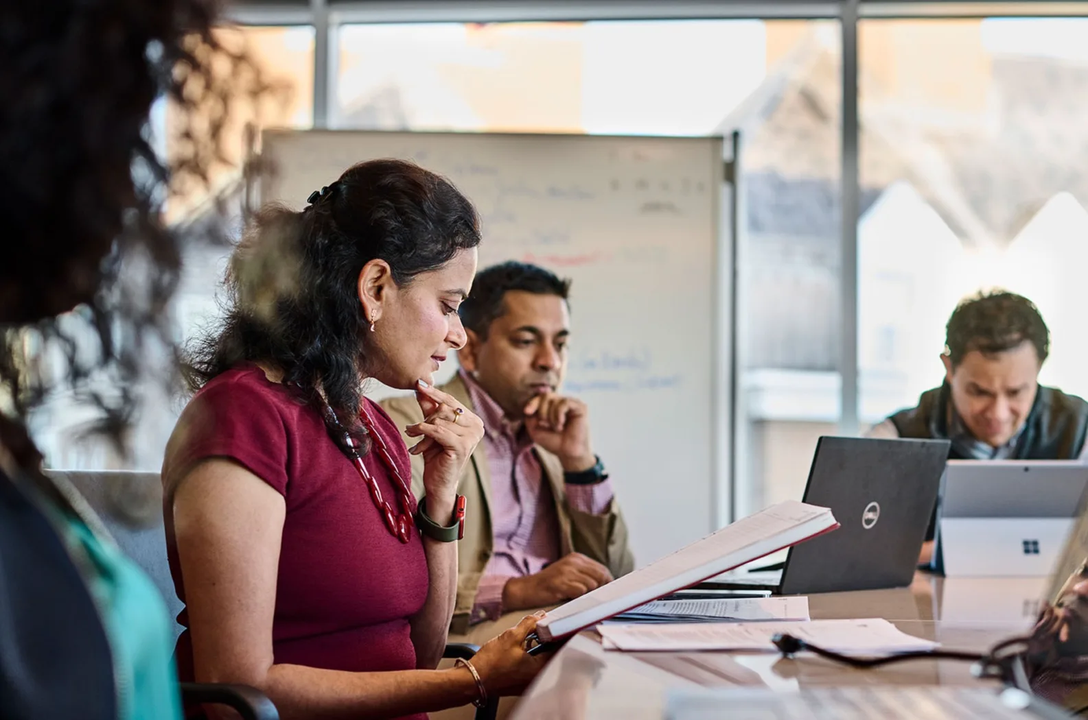 Male and female executives review their notes on paper and on their laptops while sitting down inside a meeting room with a white board in the background