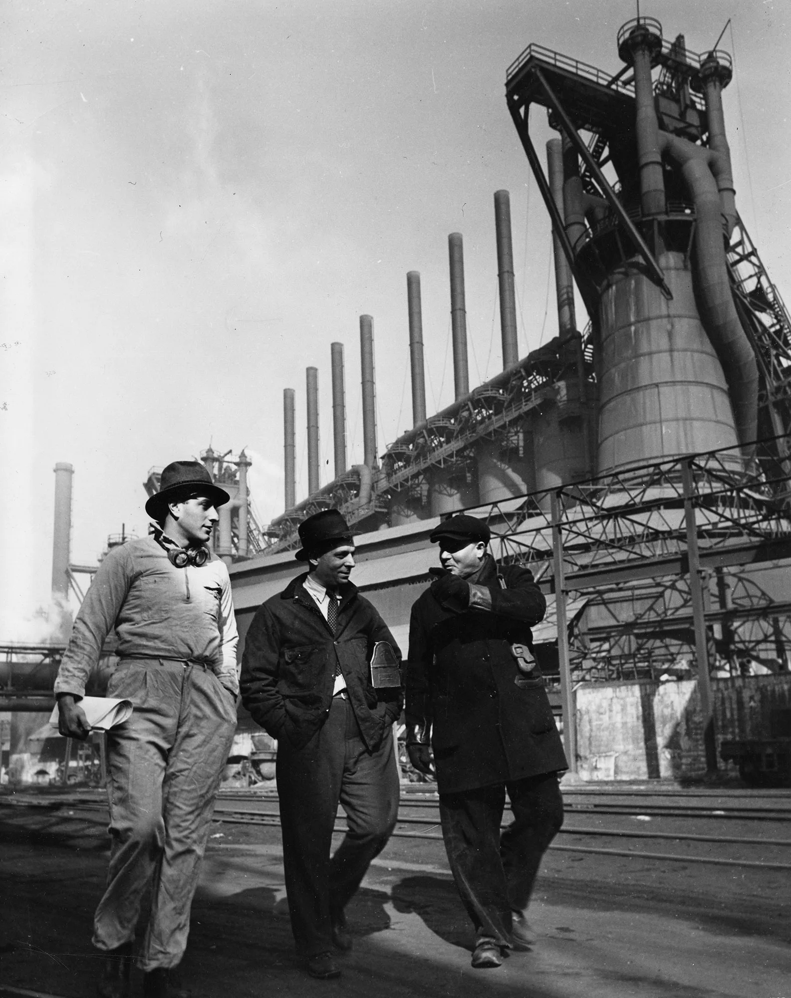 Three industrial workers talk while walking outside a steel mill with tall smokestacks.