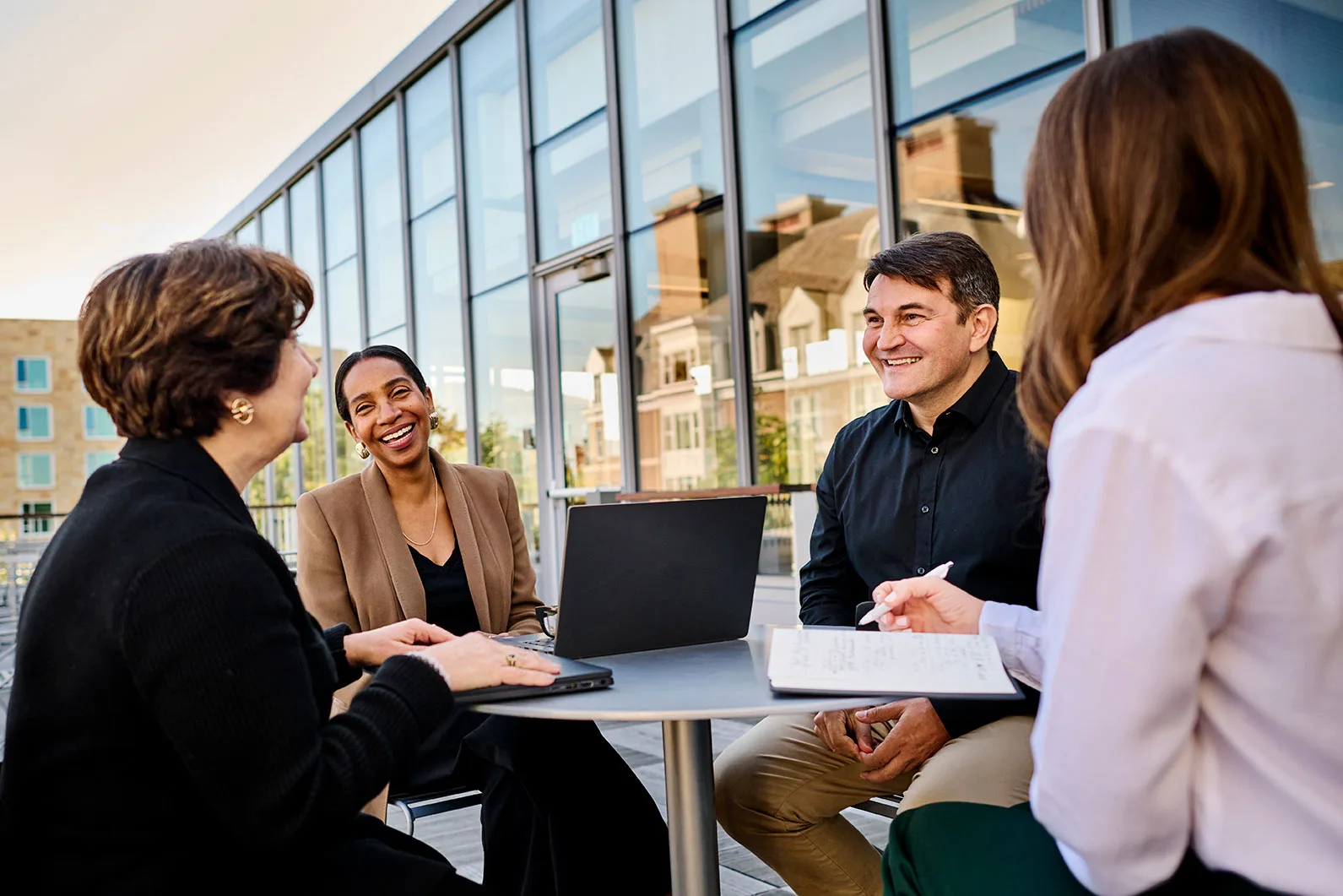 Executives gather around a table outside, engaged in a discussion around a laptop  and share a laugh together.