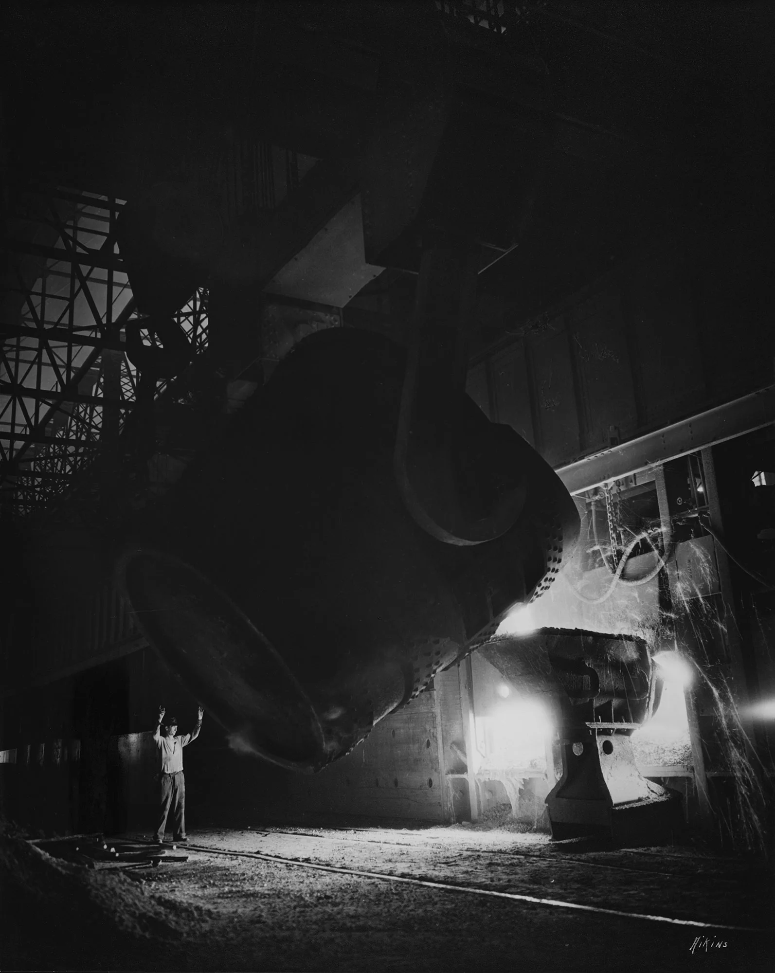 A large steel ladle pours molten metal into a mold as a person stands nearby guiding the process in a dark industrial foundry.