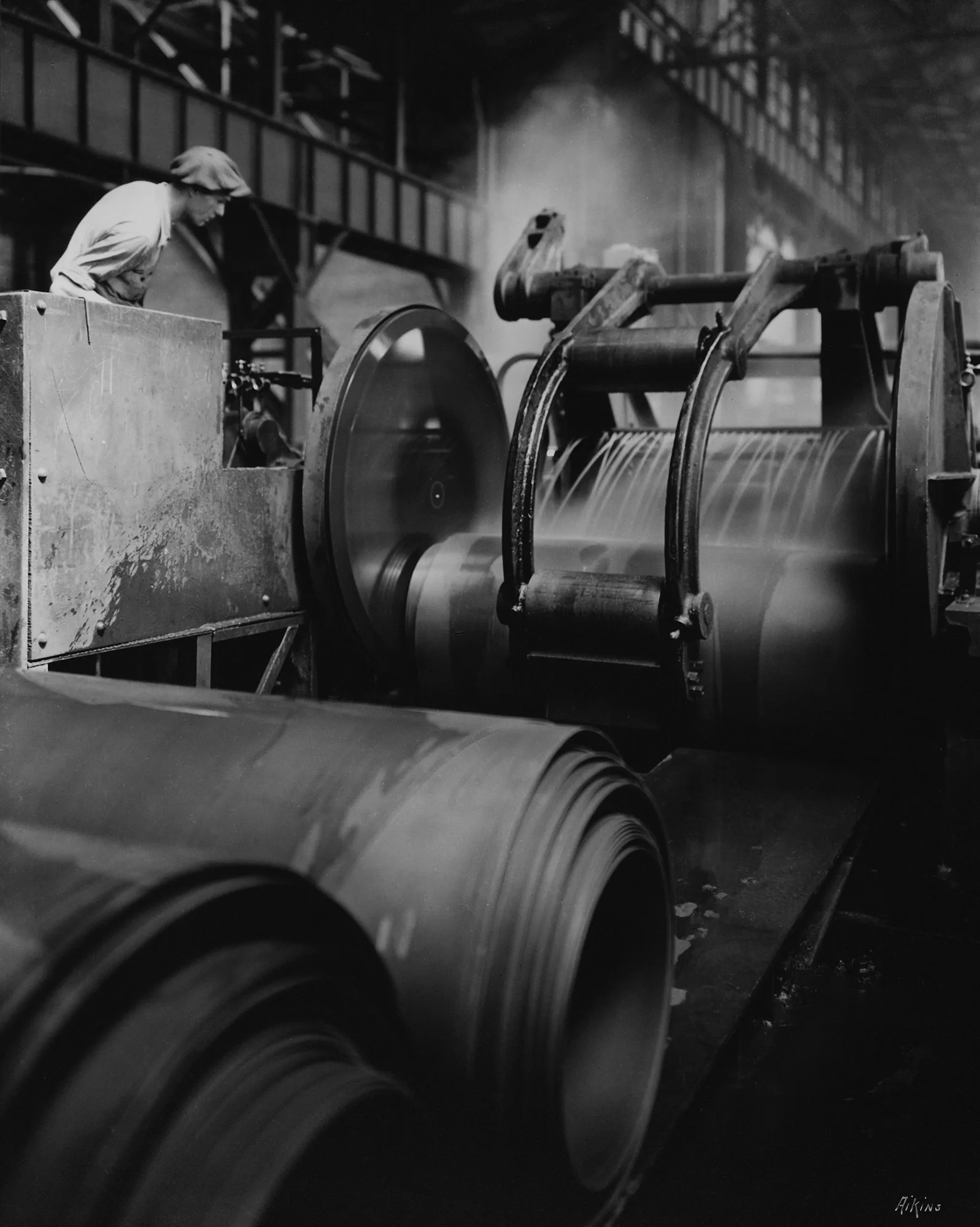 Worker monitors large industrial rollers processing hot steel sheets in a steel mill with steam rising in the background.