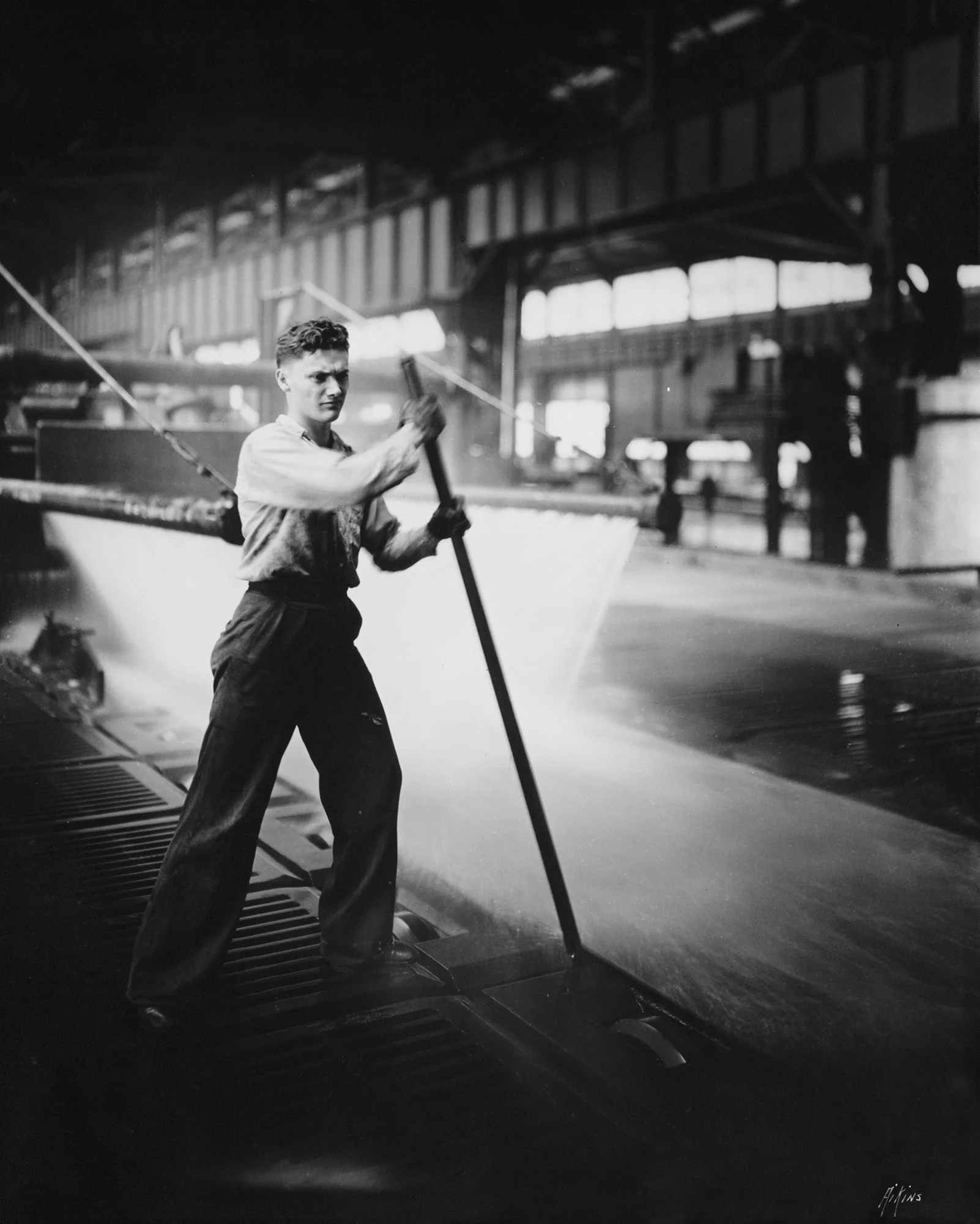 Individual uses a long pole to guide steel under a water spray cooling system inside a large industrial mill.