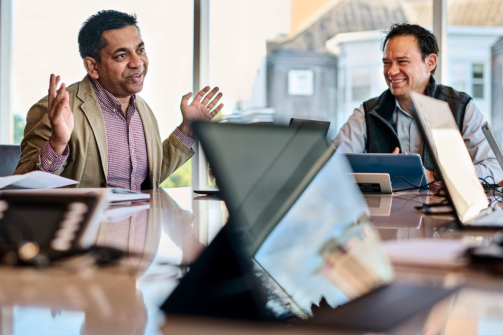 Two male executives sit at a conference room table with open laptops and talk