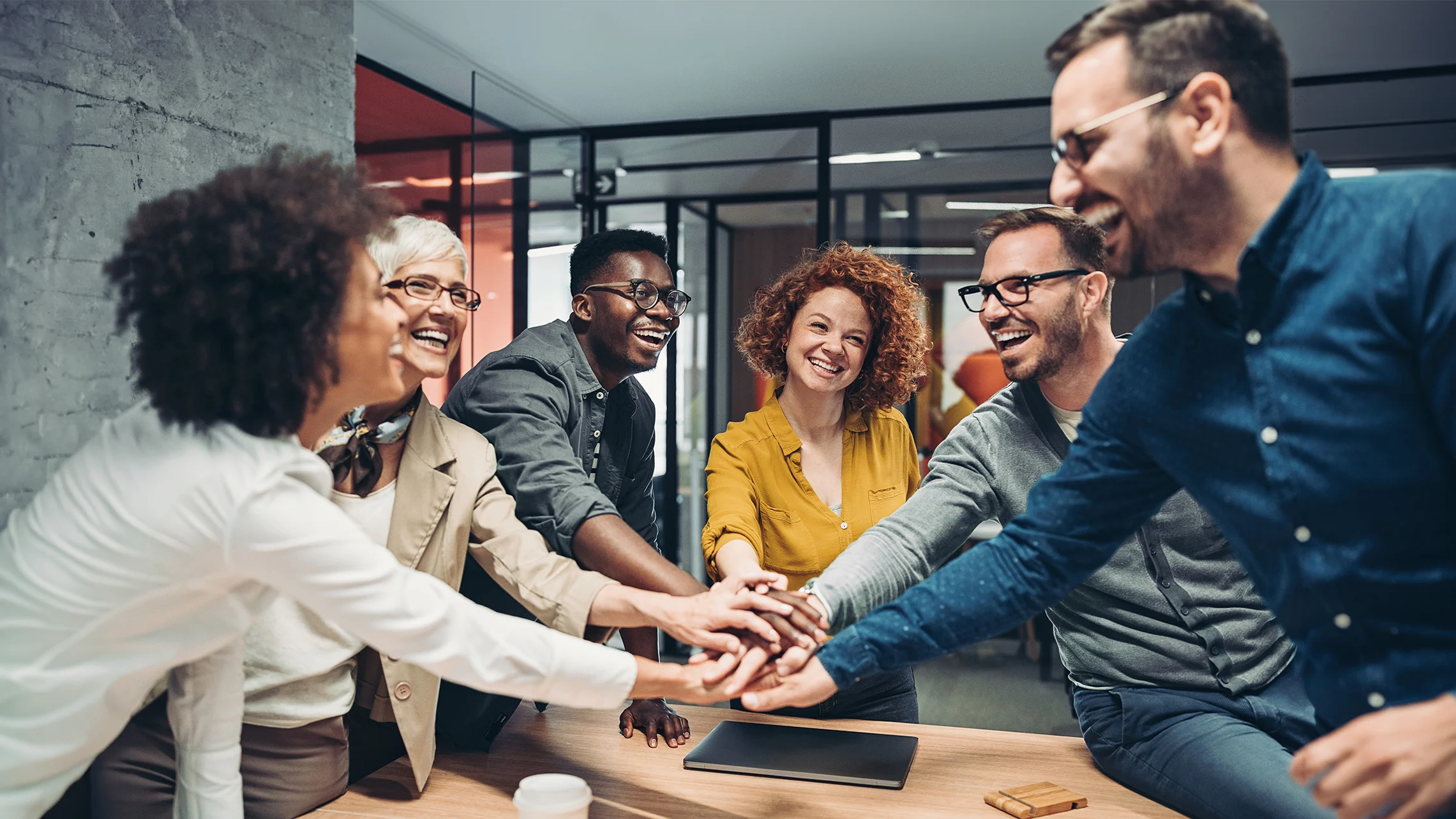 Men and women of various ages gather around a table and stack their hands in the center to show unity