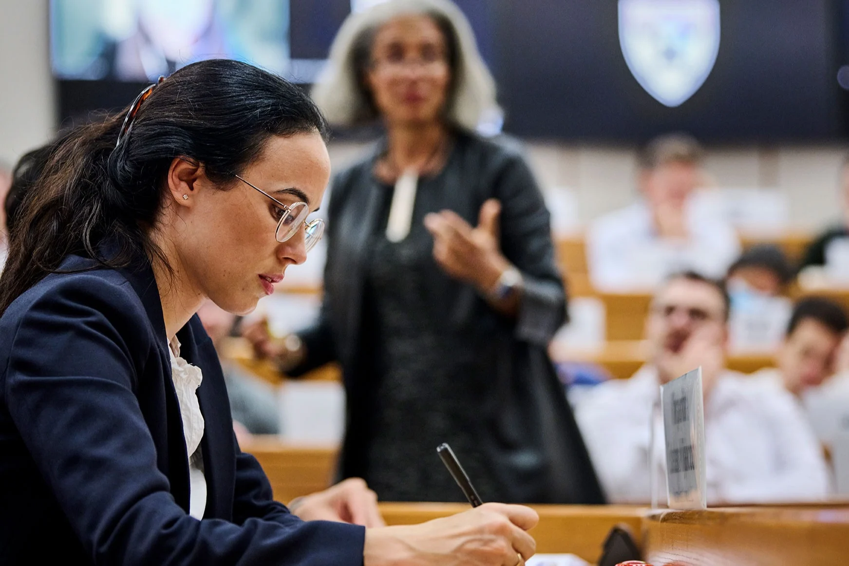 A female participant takes notes inside the classroom as the professor lectures in the background