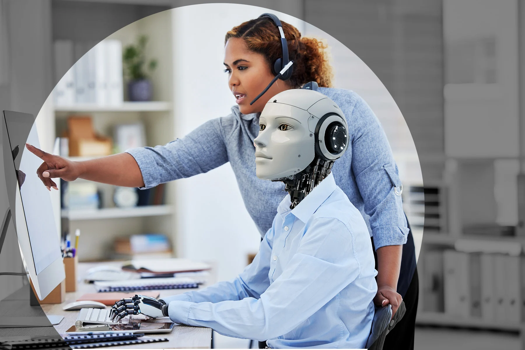 Person standing pointing out something on computer screen to robot sitting at desk
