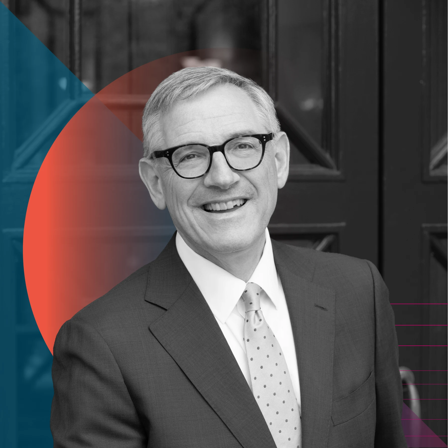 A black and white portrait of a smiling person with short hair, wearing glasses, a dark suit, and a light polka-dot tie, standing in front of dark double doors with a blue and red geometric overlay.