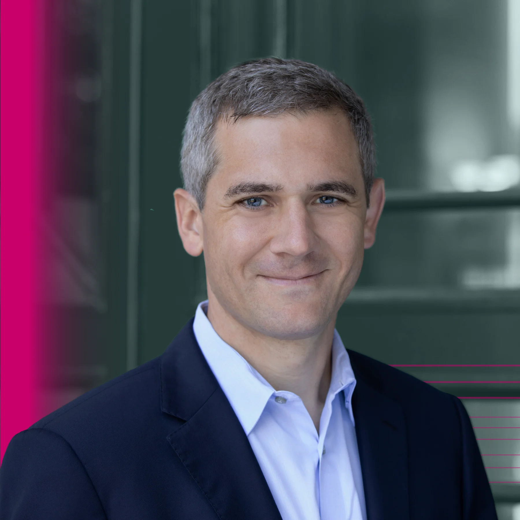 Portrait of a man with short gray hair wearing a dark blazer and light-colored dress shirt. He faces the camera with a slight smile. The background is softly blurred, with green framing and pink graphic accents along the left and lower right edges.