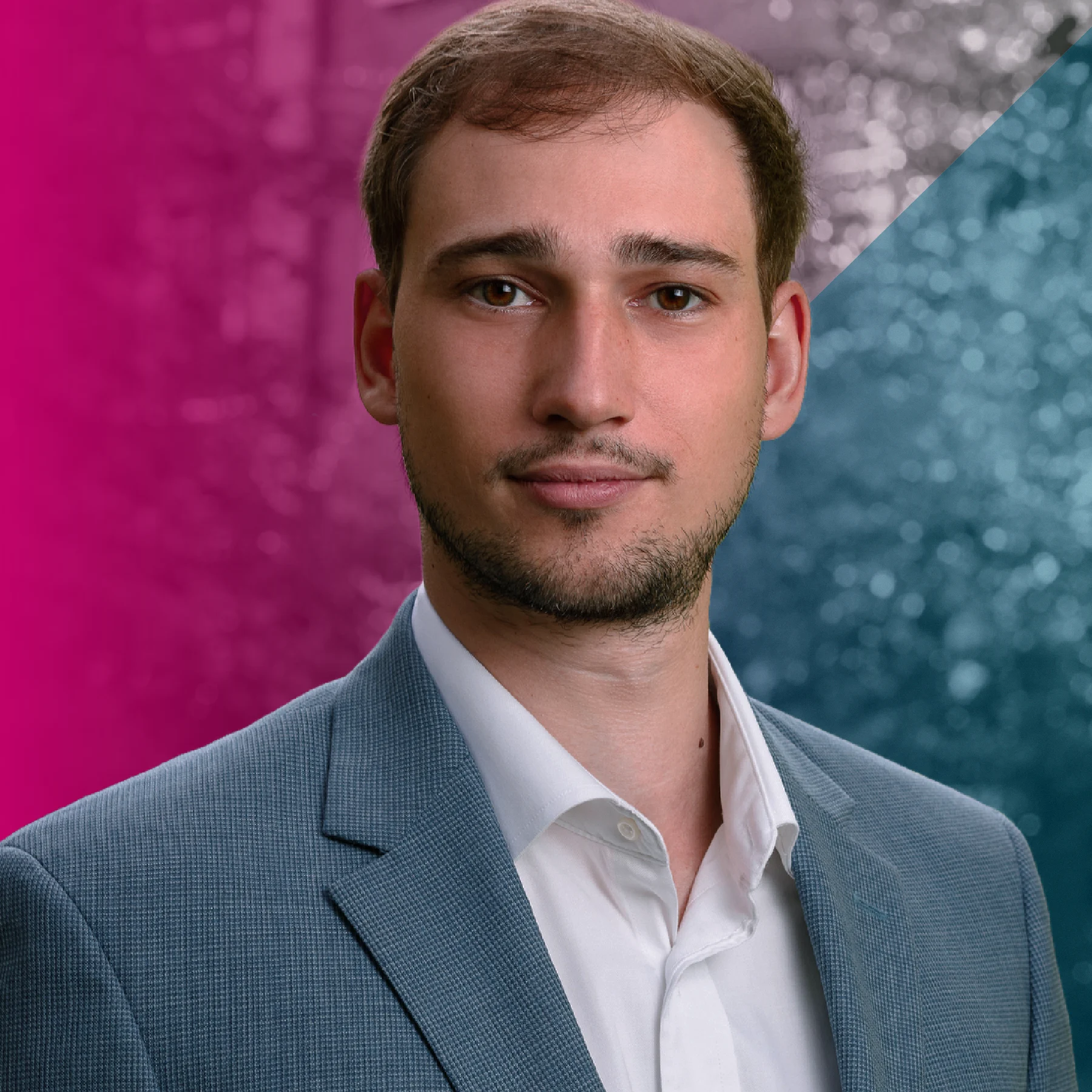 A person with short brown hair and trimmed facial hair faces the camera, wearing a light blue blazer and white collared shirt. The background is blurred with pink tones on the left and blue tones on the right.