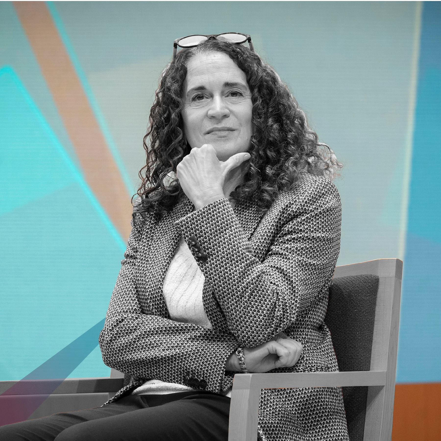 A woman with curly hair sits in a chair, facing the camera. One hand rests under her chin. She wears glasses on her head, a patterned blazer, and a light sweater. The background shows soft shapes in blue and orange. The woman is in black and white.