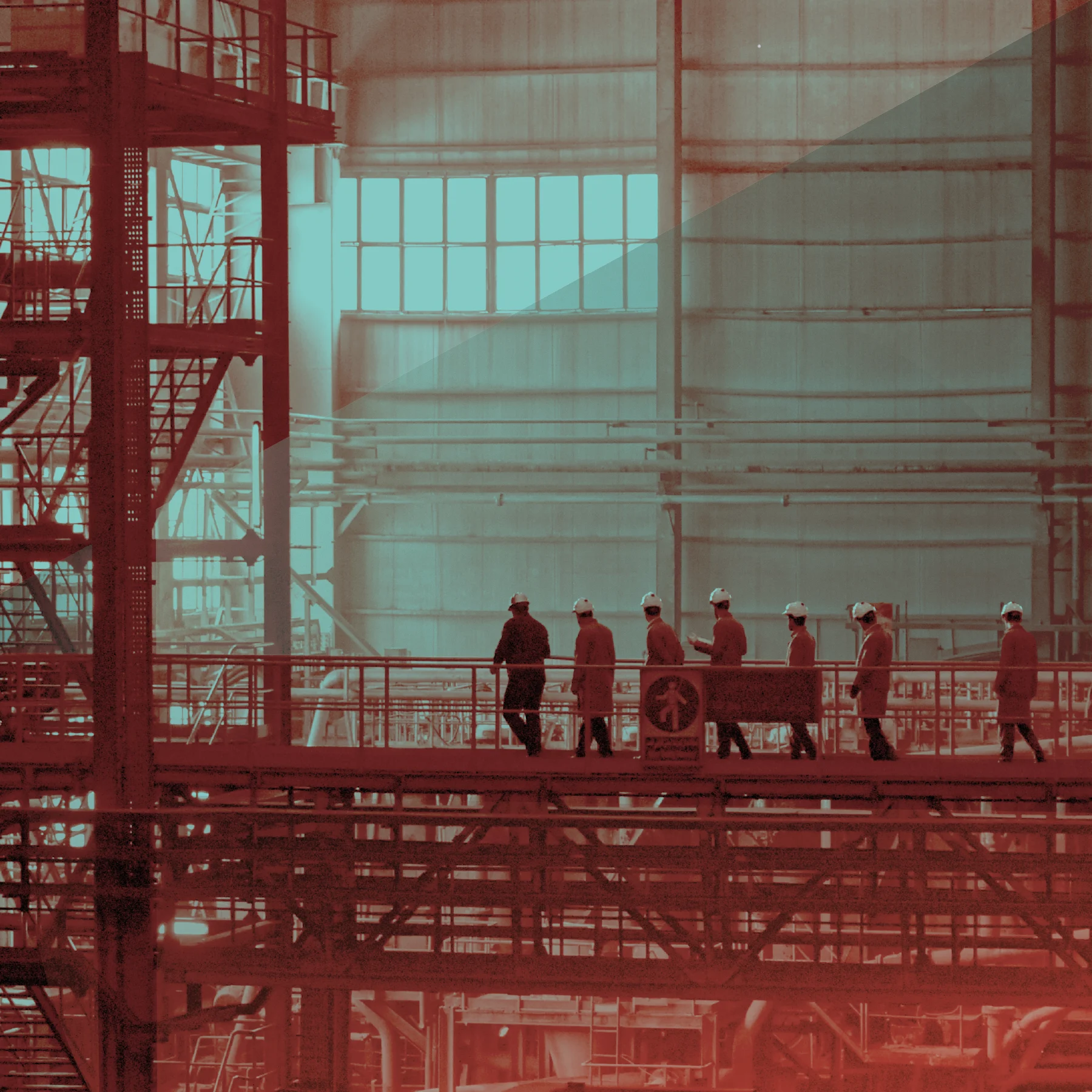 Workers in hard hats walk across an elevated platform inside a large industrial facility with metal beams, pipes, and tall windows, tinted in red and teal.