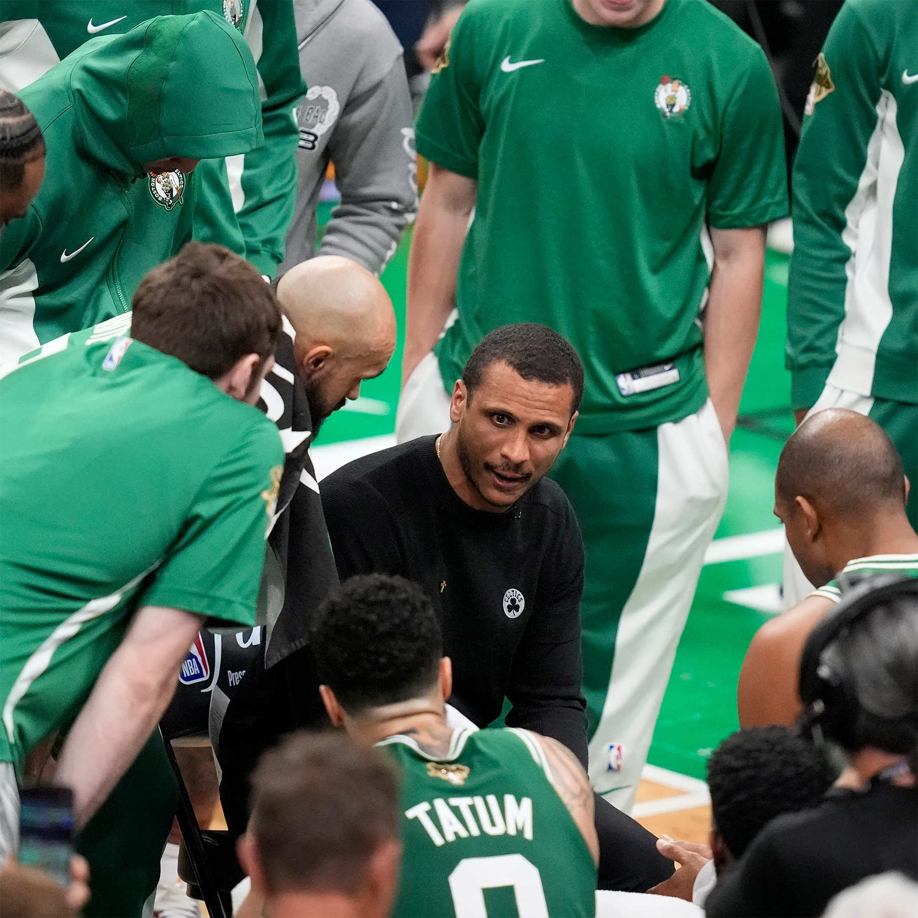 Joe Mazzulla, in a black shirt is seated, surrounded by men in green and white basketball uniforms.