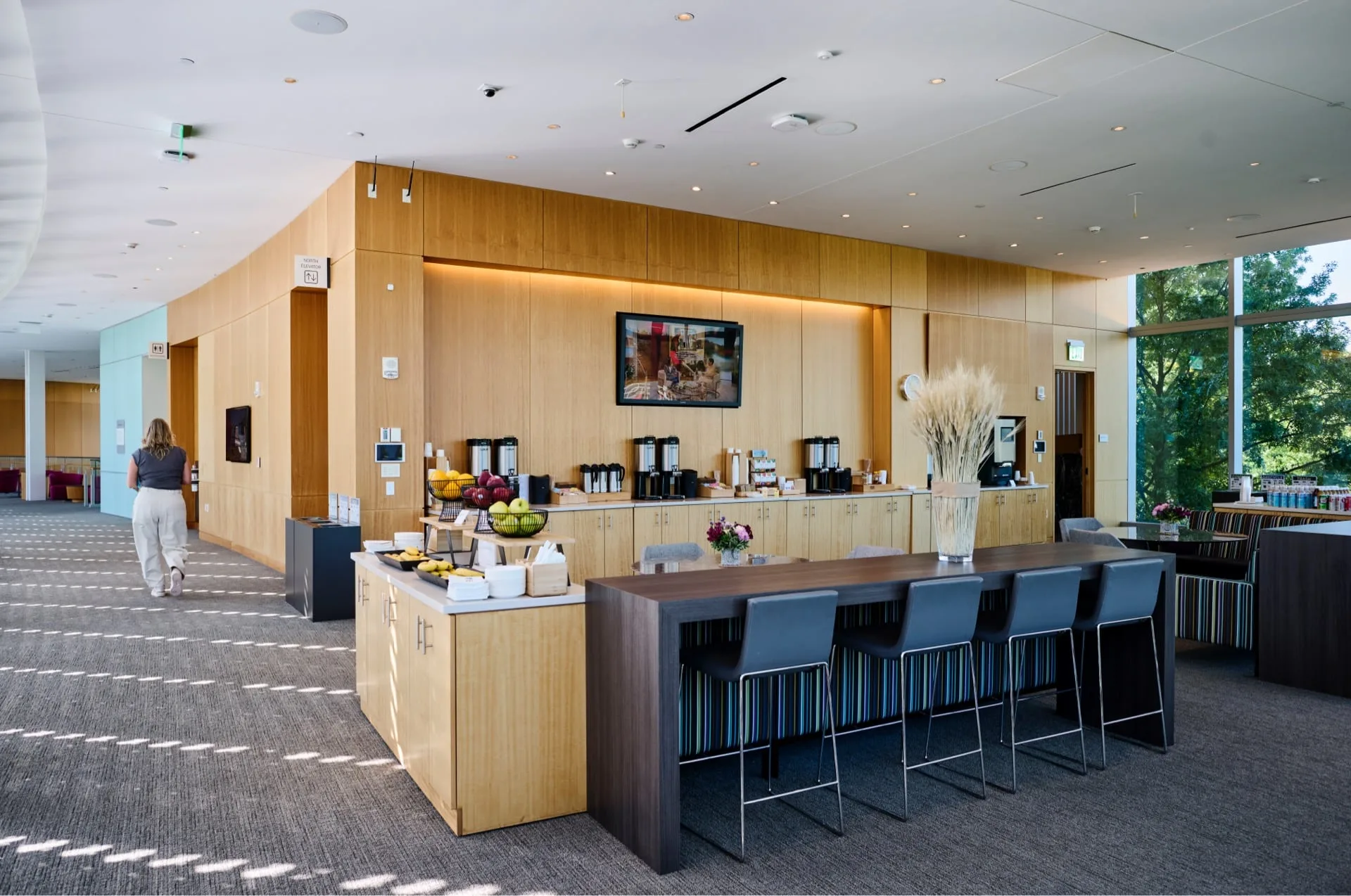 Modern lounge in Tata with wood paneling, coffee station, and fruit display. Large windows let in natural light, creating an inviting space