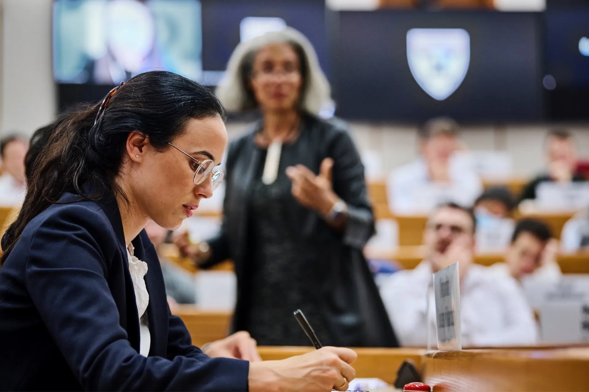 A female participant takes notes inside the classroom as the professor lectures in the background