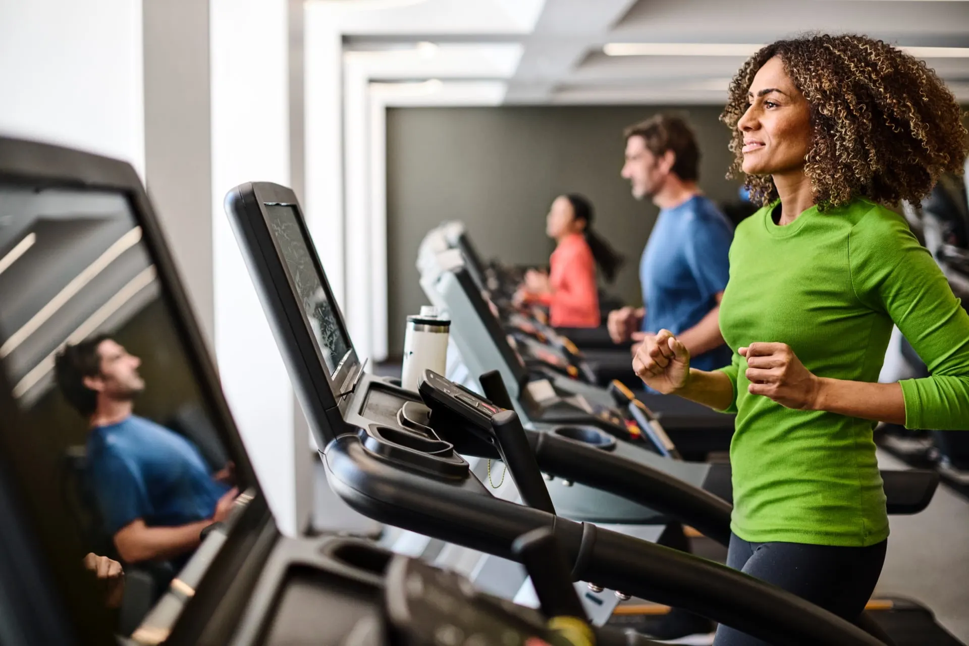 Smiling woman in a green shirt runs on a treadmill in Shad gym, with other people exercising in the background 