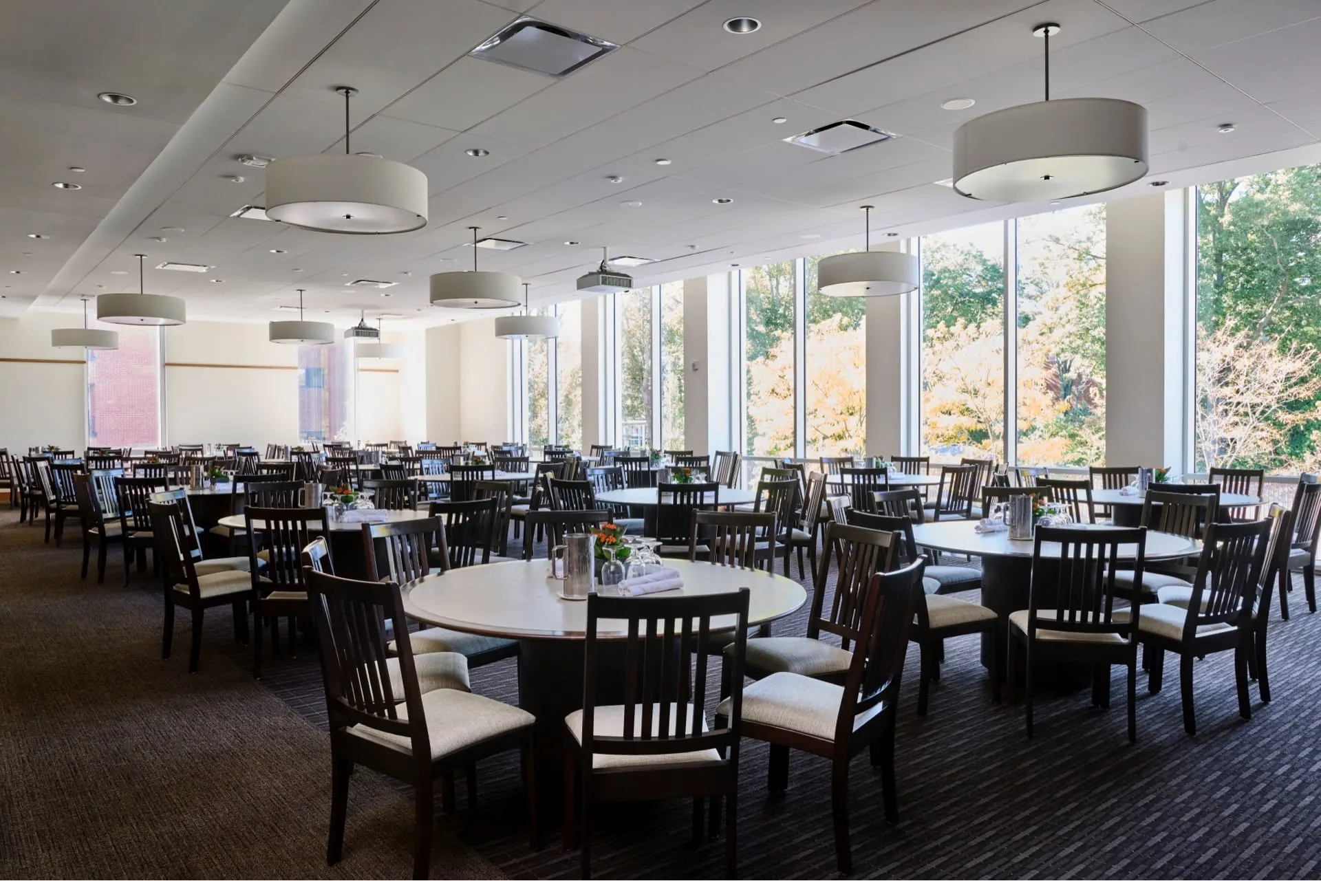 A spacious, well-lit dining Chao Dining hall with round tables and wooden chairs, set with tableware, overlooking trees through large windows