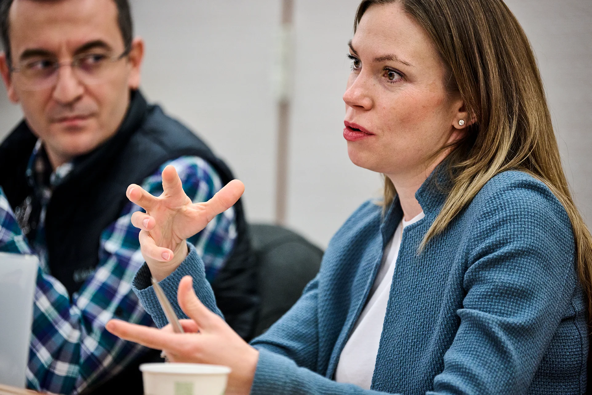 Close up of a woman in a blue blazer speaking in a classroom. A man in a plaid shirt and black vest with glasses listens attentively