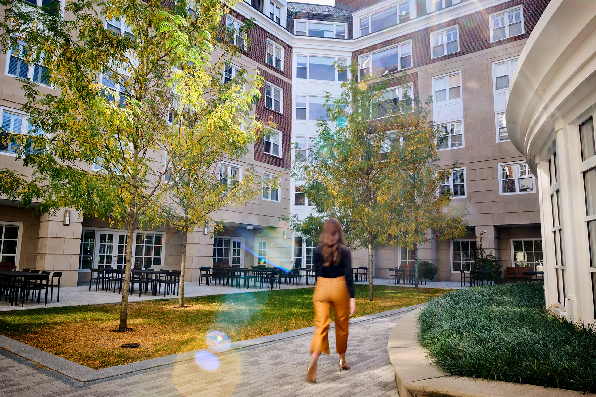 A woman in stylish attire walks through a sunlit McArthur courtyard surrounded by modern brick buildings and autumn tree