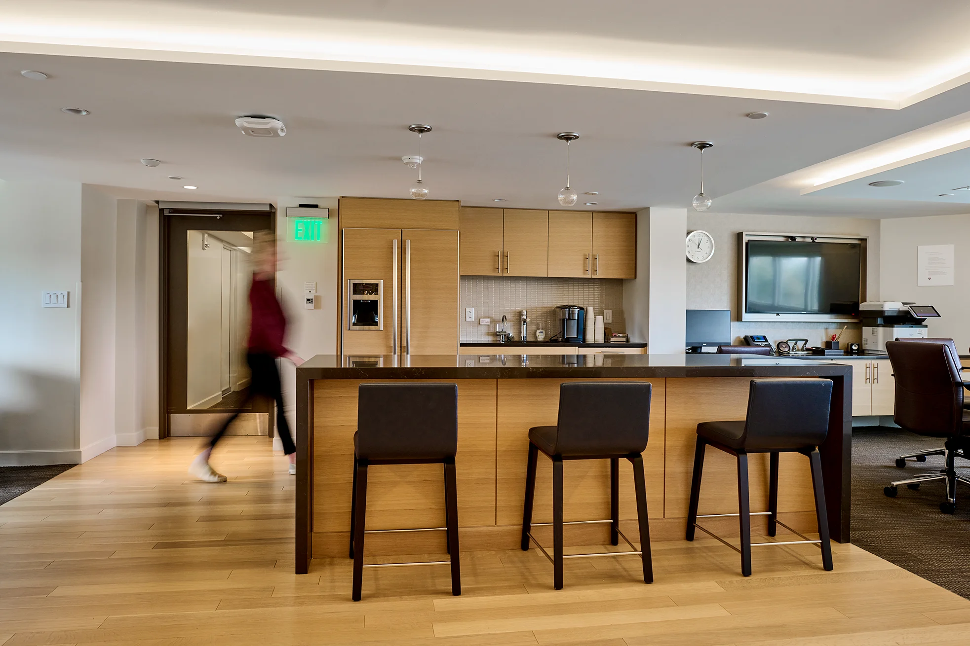A modern kitchen area with wooden cabinets, bar stools, pendant lights, and a blurred figure walking by