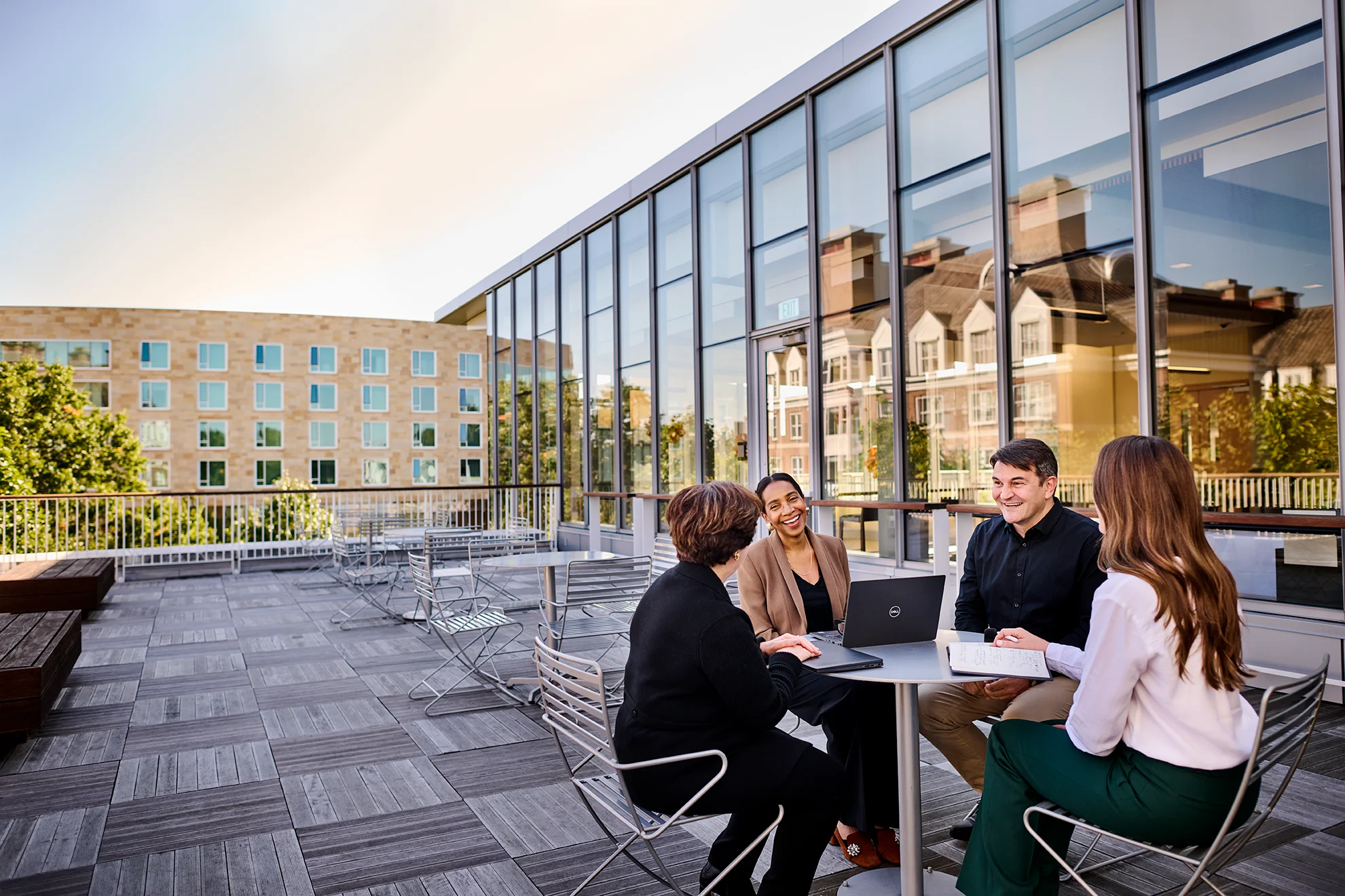 Four participants engages in a discussion at Chao Center outdoor café, surrounded by modern glass architecture reflecting HBS campus buildings