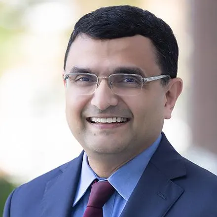 V.G. Narayanan wearing glasses, a navy suit, a light blue shirt, and a burgundy tie. Blurred outdoor background.