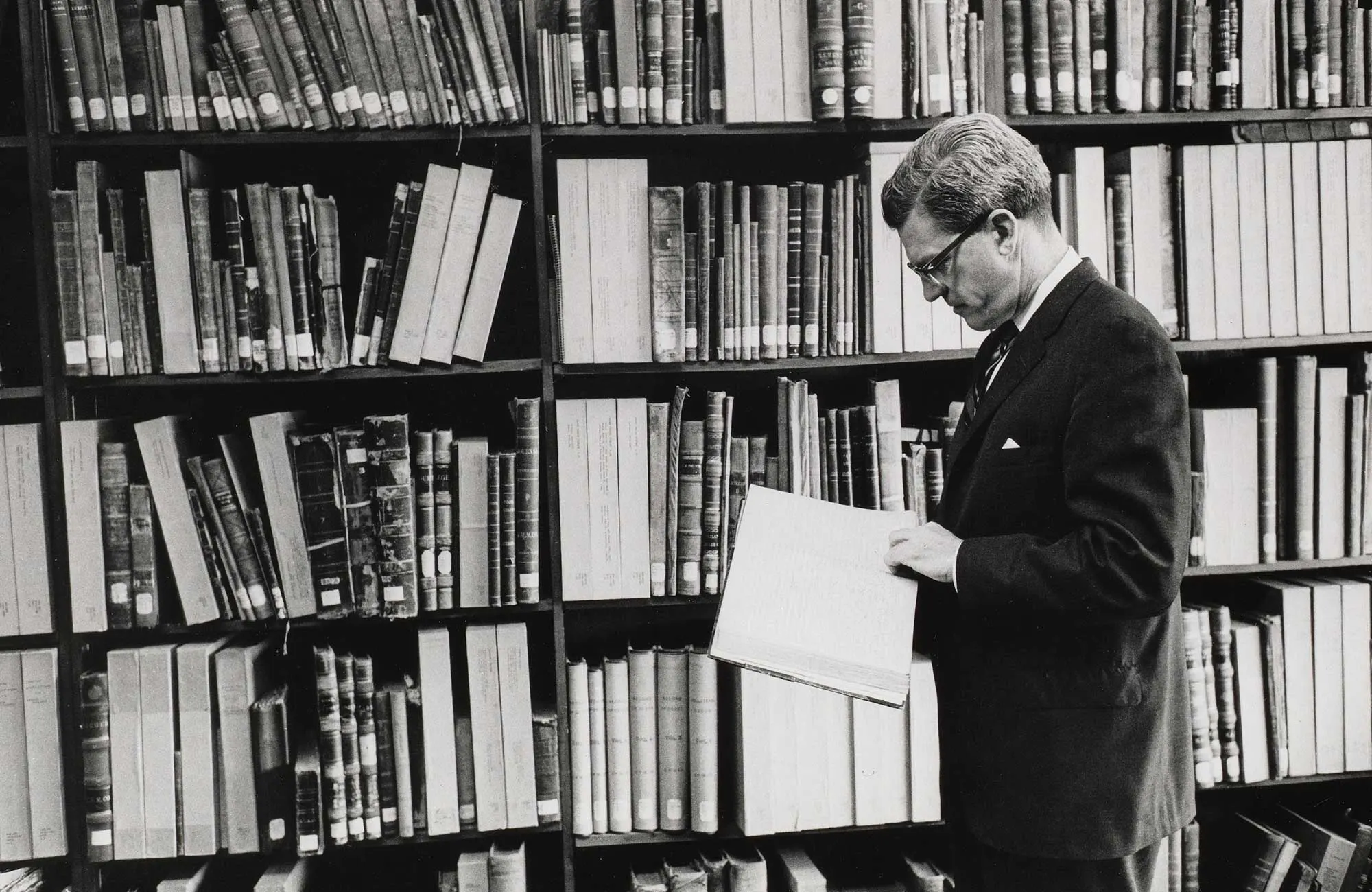 A man stands in front of a tall shelf packed with books, some with their spines facing outward and many with their pages facing out. He looks down at an open book in his hands.