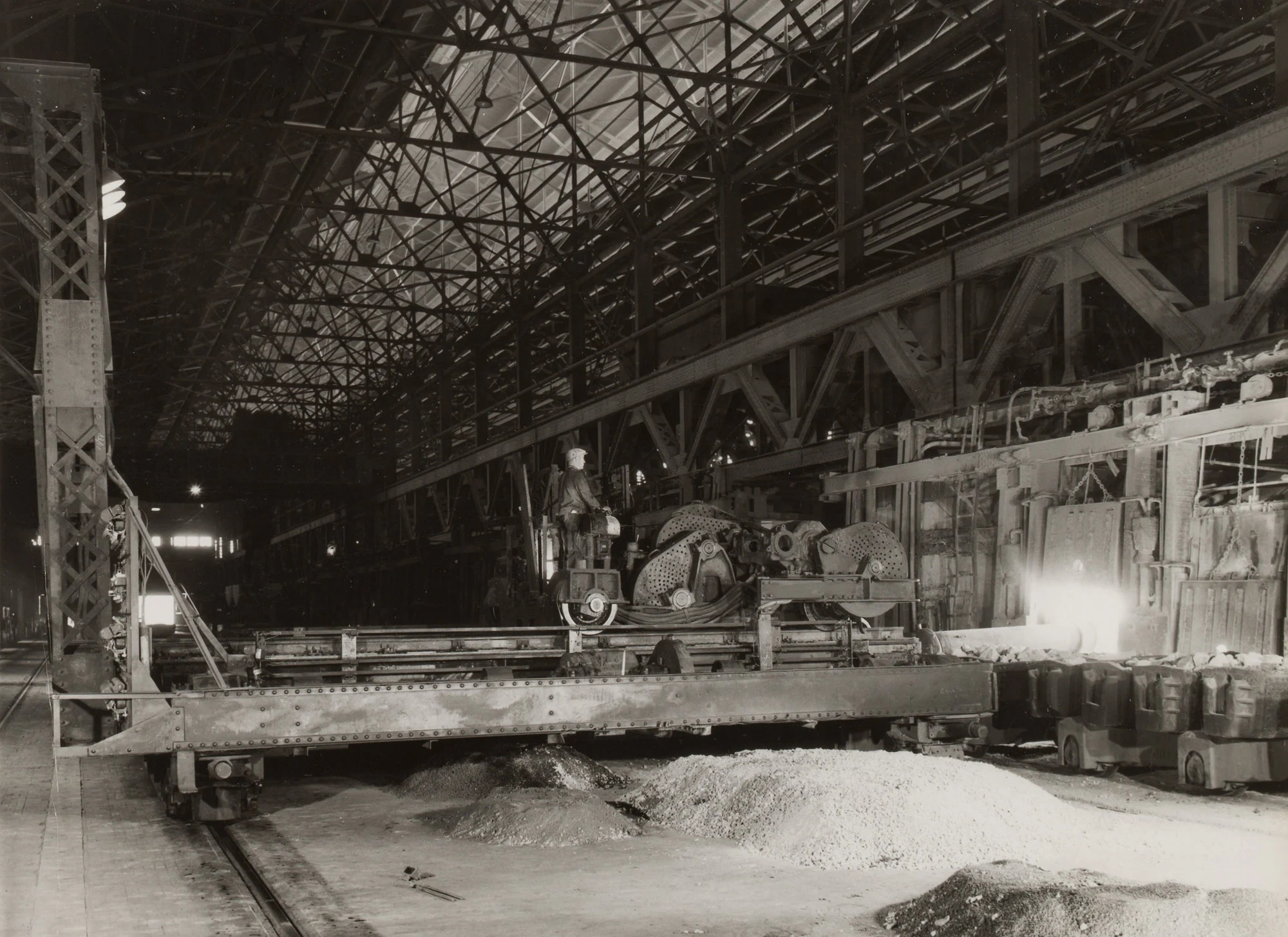 Steelworker operates machinery inside a large mill as molten metal pours into molds under a truss-supported industrial roof.