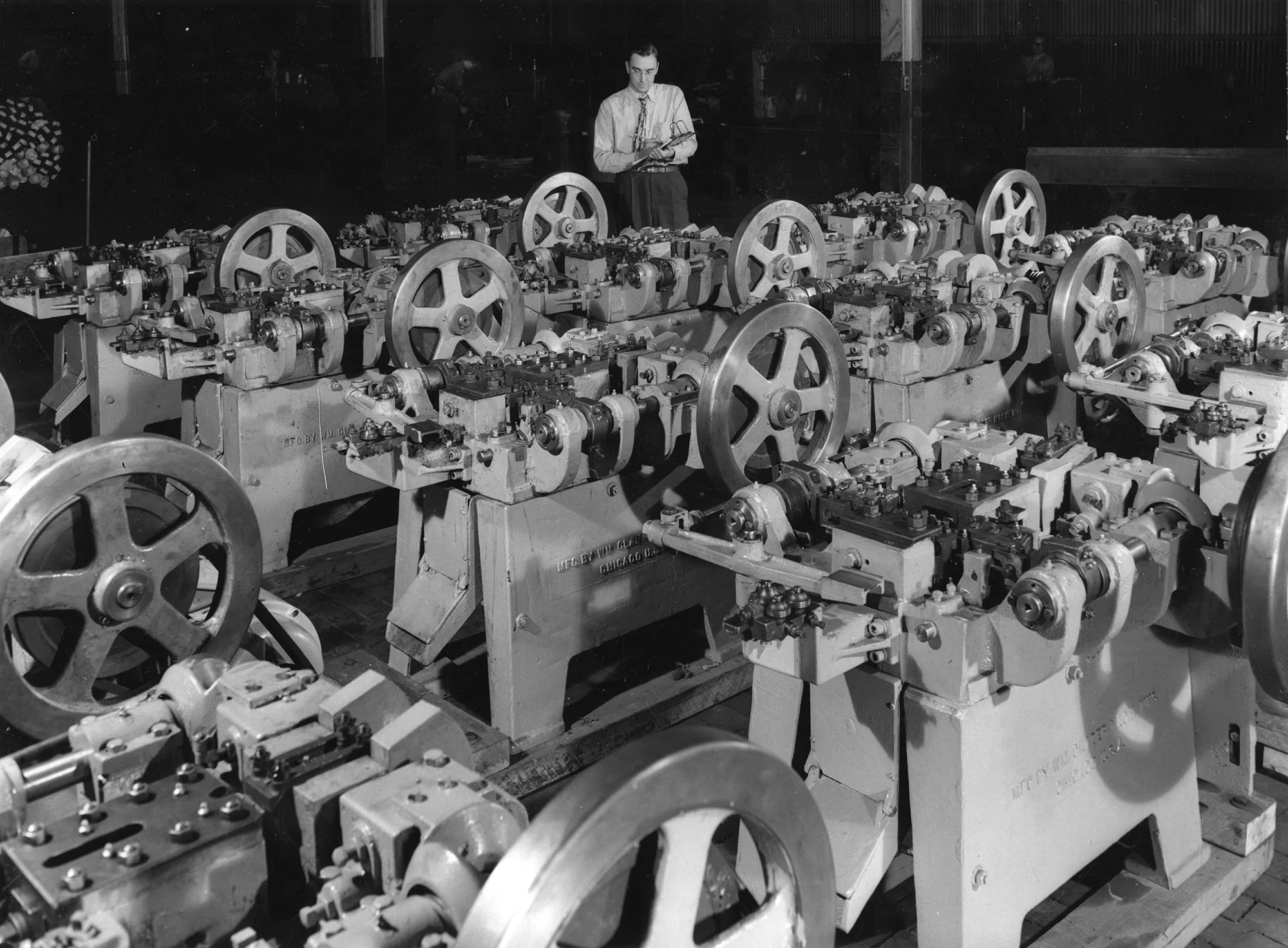 Individual inspects rows of identical industrial metalworking machines with large flywheels in a factory setting.