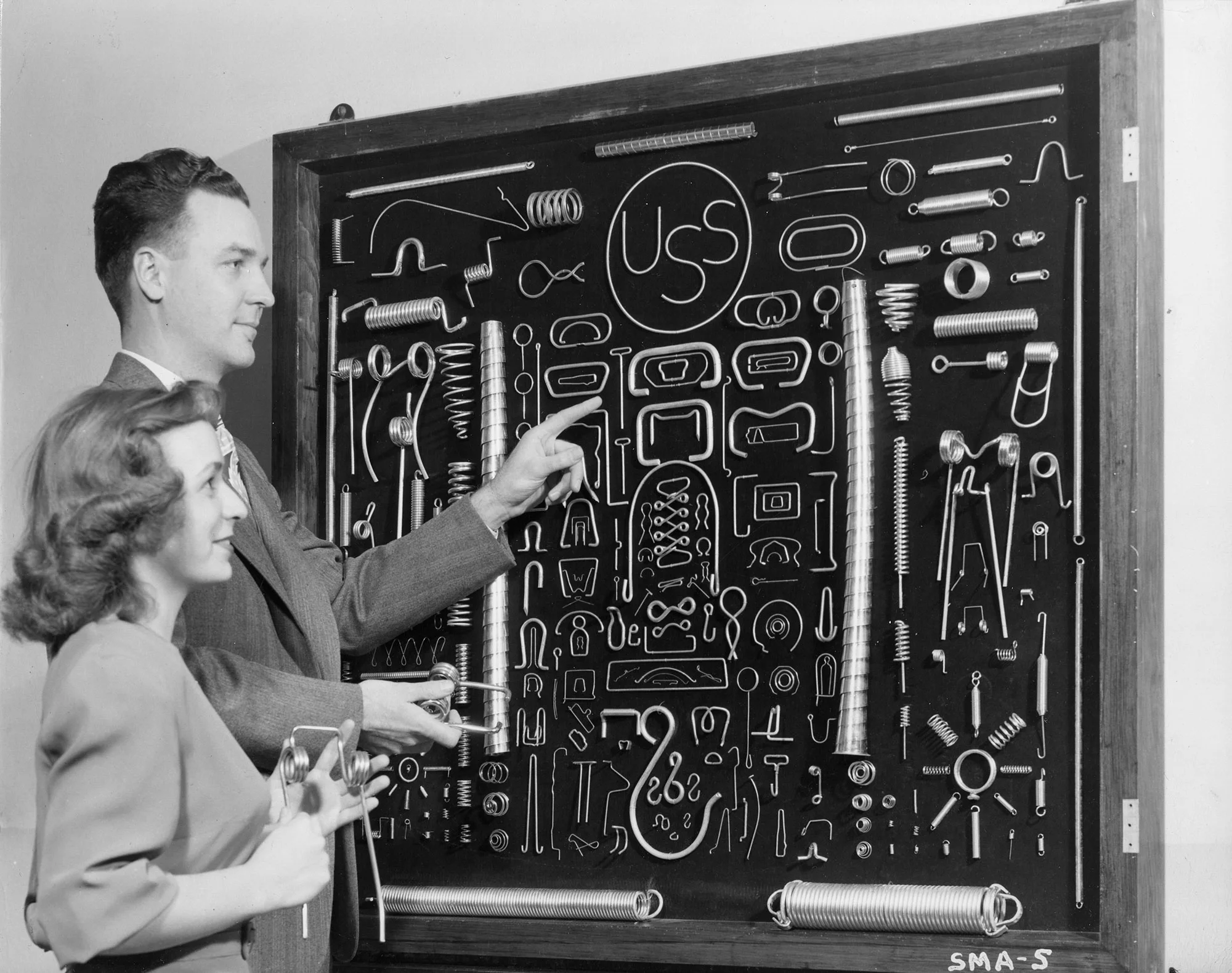 Two individuals examine a display board of various metal springs and wire forms labeled with the USS logo.