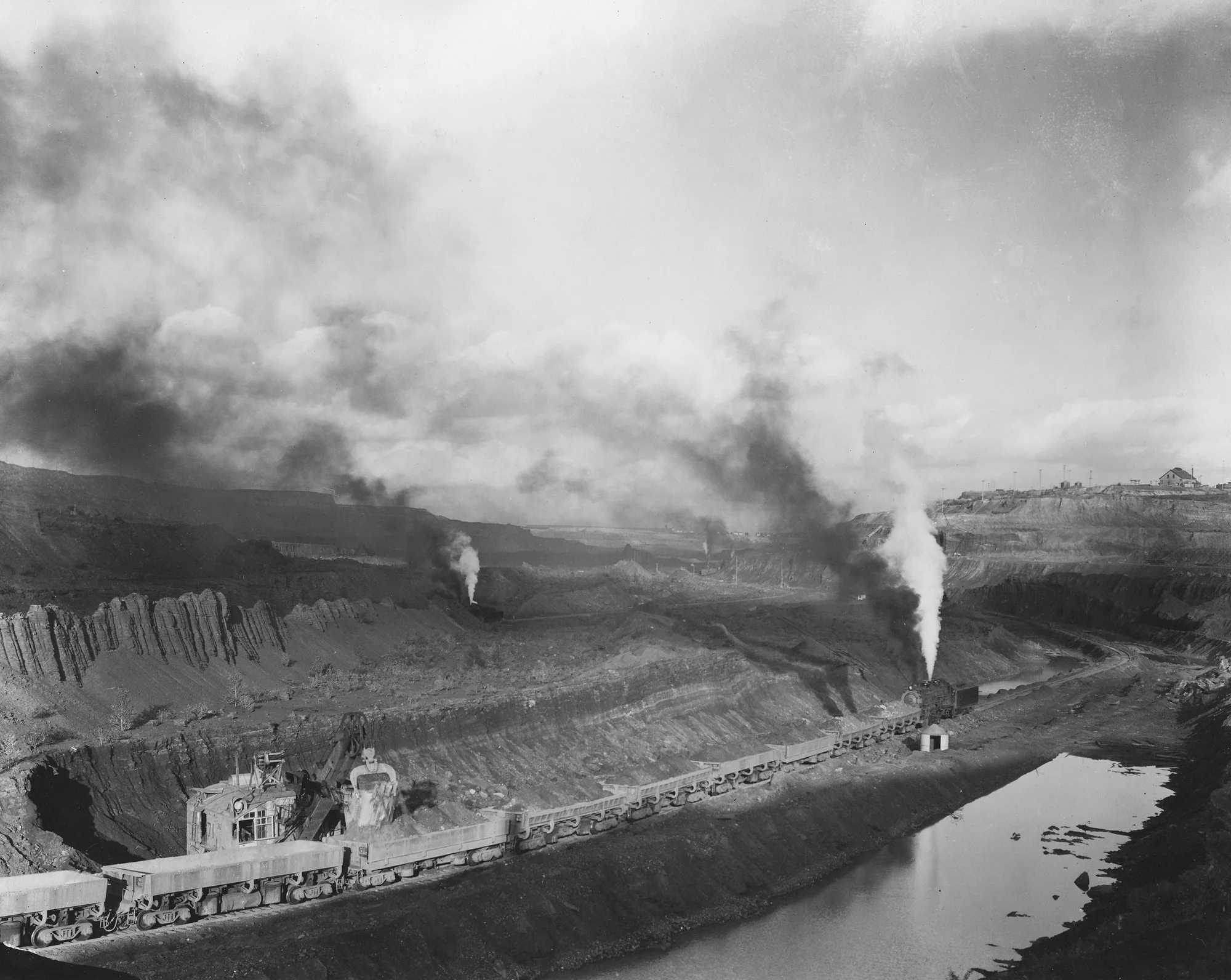 Open-pit iron ore mine with steam-powered shovels loading railcars and smoke rising across a wide, industrial landscape.