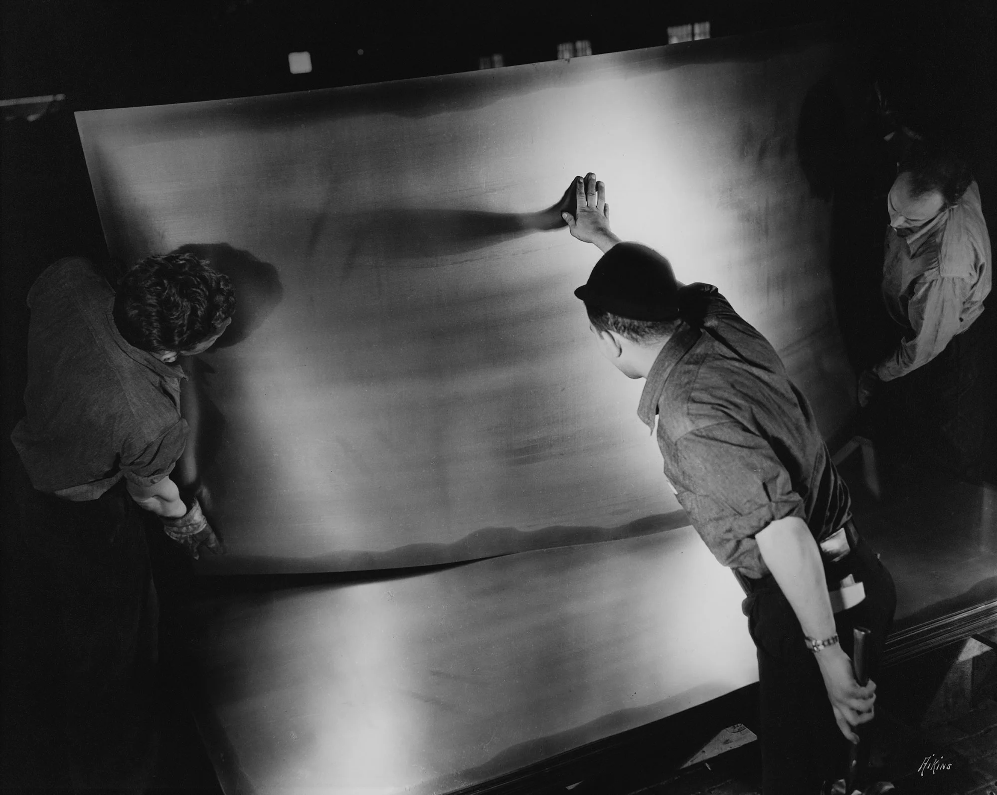 Three individuals inspect a large, reflective steel sheet under bright industrial lighting in a factory setting.